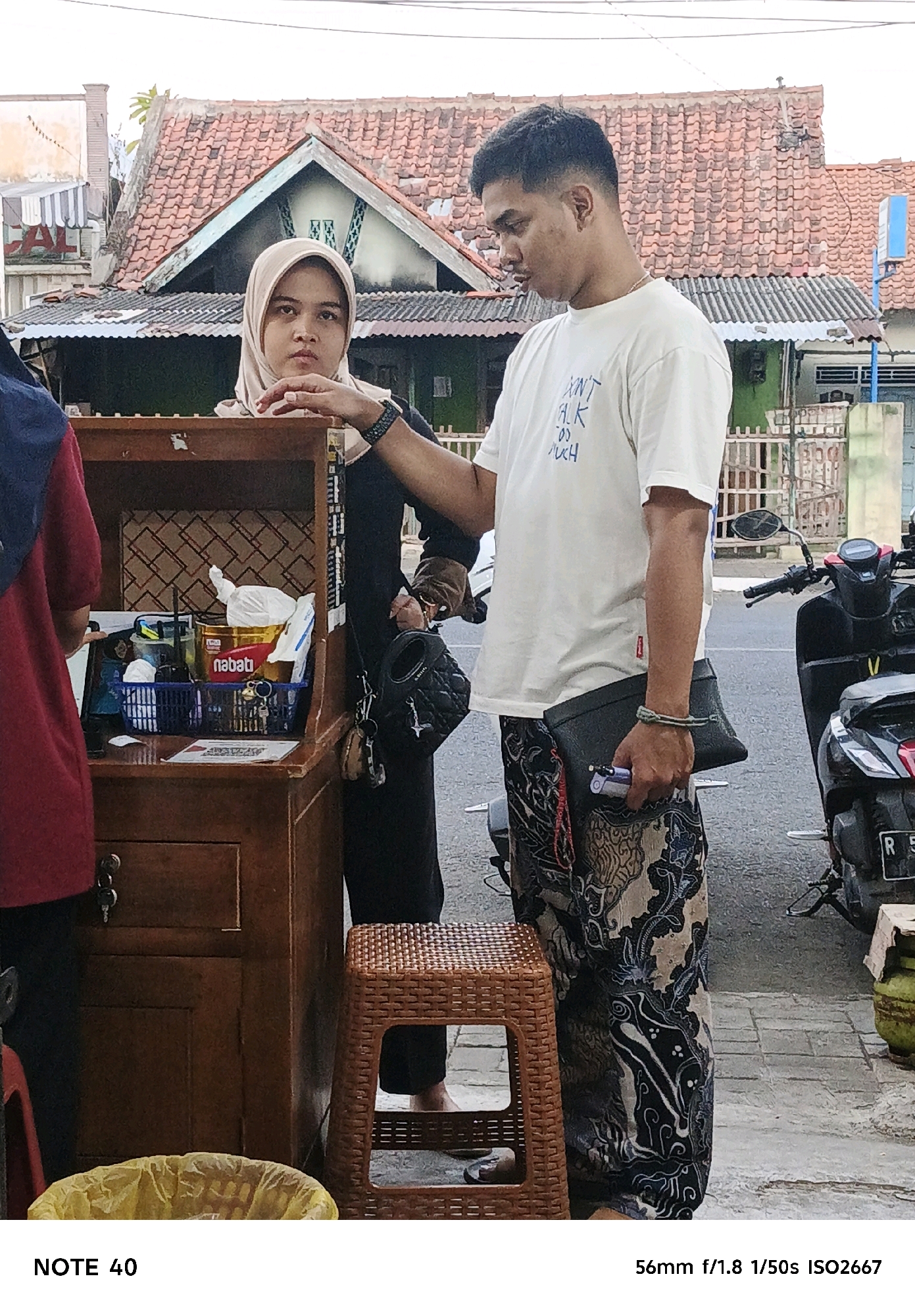 A man and a woman stand near a wooden cart on a sidewalk. The woman, wearing a light-colored hijab and black clothing, faces the camera with a neutral expression. The man, in a white t-shirt and patterned pants, looks down and to the left, holding a black clutch bag and a vape. The cart contains various items, including a red and gold cylindrical tin labeled "nabati," a blue basket with white items, and a yellow plastic bag visible at the bottom of the frame. In the background, a tiled roof house and a parked motorcycle are visible. The scene appears to be in a commercial or residential area, possibly in Cilacap, Indonesia, given the "NOTE 40" text which may refer to a location or item.