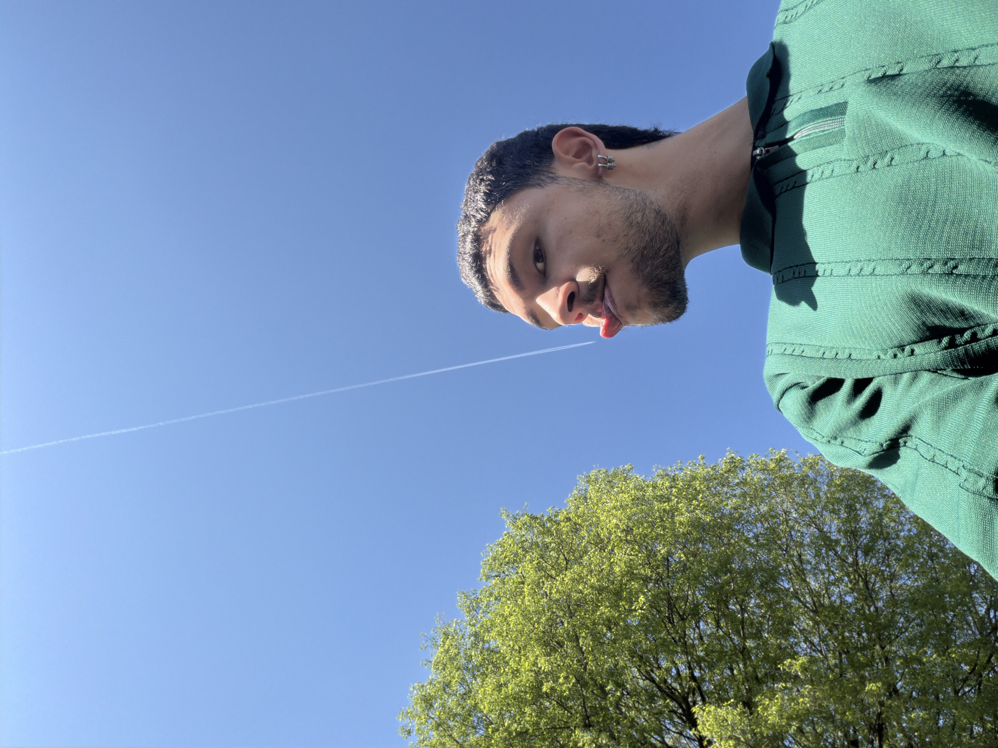 A low-angle shot from outdoors shows a light-skinned person wearing a green collared shirt. The person has short dark hair, a beard, and is sticking their tongue out. In the background, there is a bright blue sky with a contrail and green trees. This scene is not identifiable to the location of Milan, Italy.