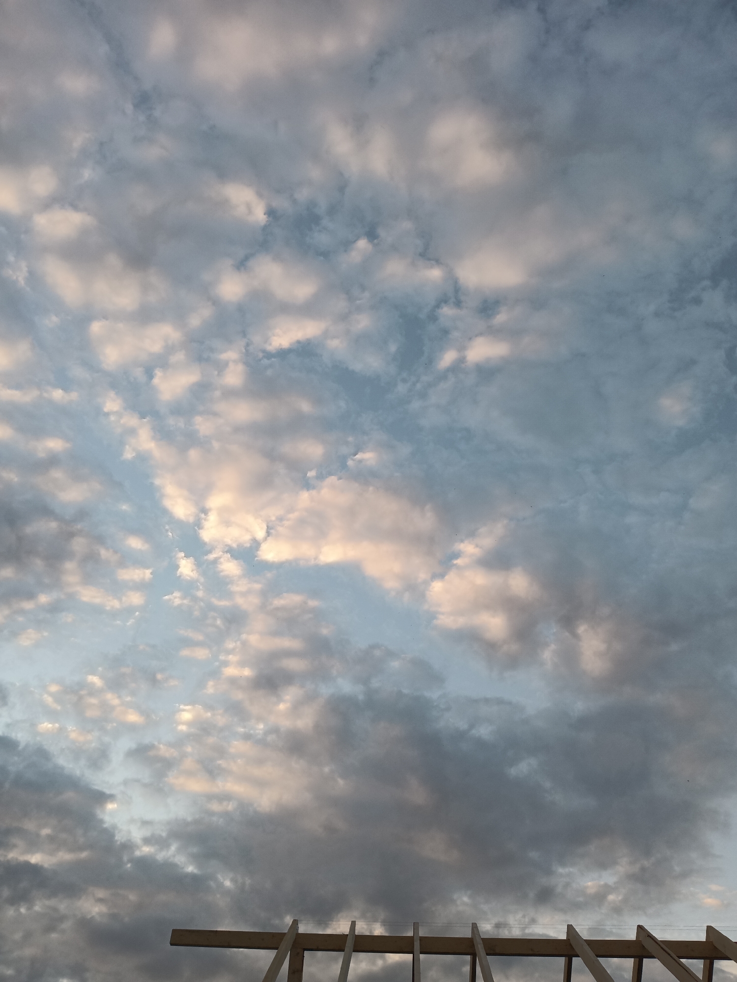 This image shows a cloudy sky over the city of Foky, Russia. The clouds are a mix of white and gray, with some patches of pink and orange light from the setting sun. The sky is a pale blue, with some areas of darker blue where the clouds are thicker. In the foreground, there is a partial view of a wooden structure, possibly a roof or scaffolding, with diagonal beams supporting horizontal ones. The scene appears to be outdoors, and there are no people visible. The overall impression is one of a serene and peaceful evening sky.