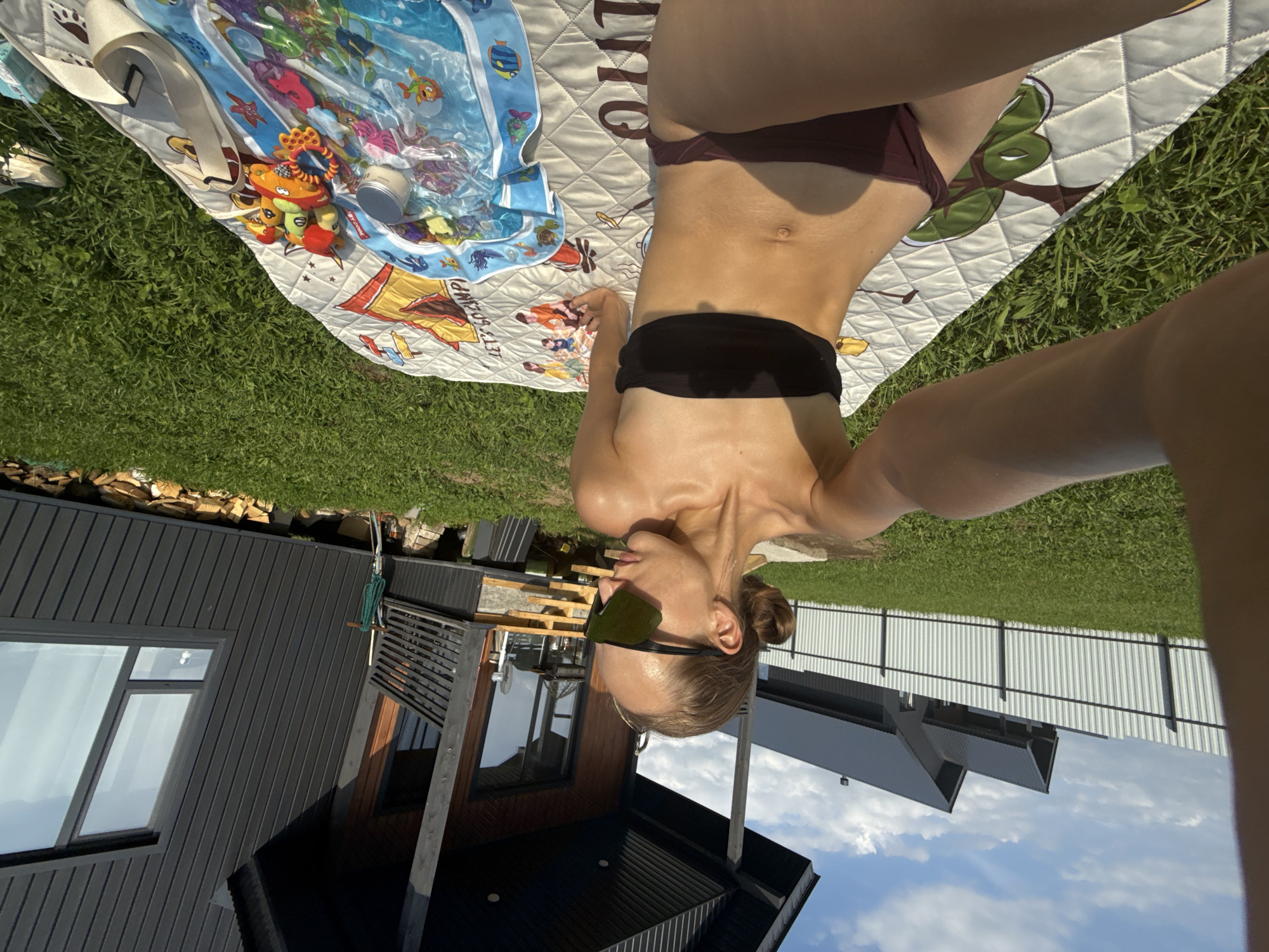 In Подшивалово, Russia, on a sunny day, a woman wearing a black bikini and large sunglasses is sitting on a patterned blanket spread on a green lawn. She is facing to her right, with her left arm extended as if taking a selfie. Next to her on the blanket are a small metal container, a brightly colored baby toy, and a blue inflatable water mat with fish designs. Behind her, a modern dark gray house with wooden accents and a sloped roof stands against a bright blue sky with scattered clouds. A white corrugated metal fence runs horizontally in the background, separating the yard from other similar houses. The overall environment is a suburban outdoor setting, likely a backyard.