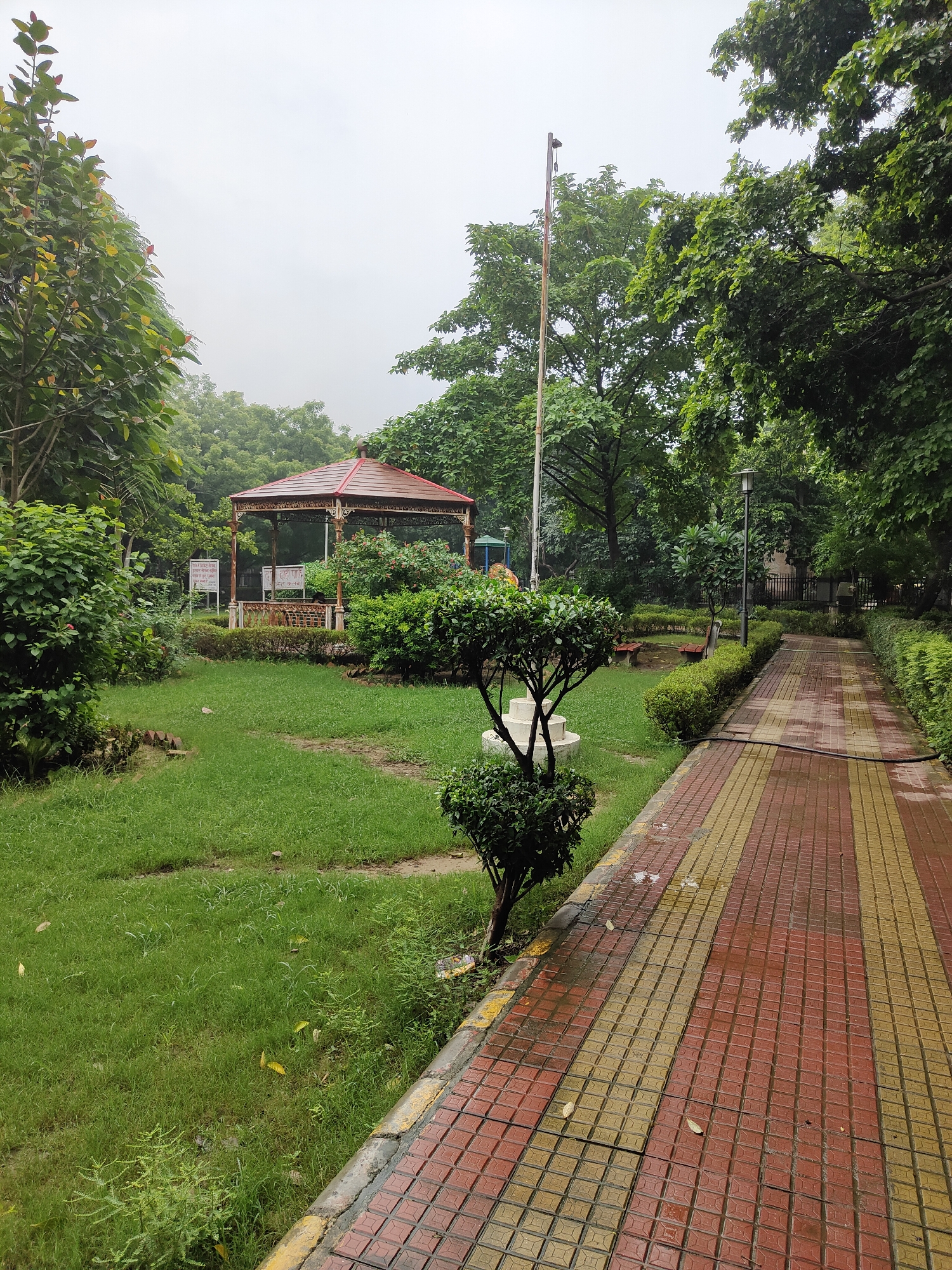 This is a landscape image of a park in Gurgaon, India. The park features a paved walkway on the right, made of red and yellow tiles, which appears wet. To the left of the walkway is a neatly trimmed green hedge. Beyond the hedge, there is a grassy area with scattered leaves and a few small trees. In the background, a red-roofed pavilion or gazebo stands among lush green trees and bushes. There are also signs visible near the pavilion. A flag pole is present to the left of the pavilion. The sky is overcast, suggesting it might have rained recently or is about to. The environment is serene and well-maintained, typical of a public park. No people are visible in the image.
