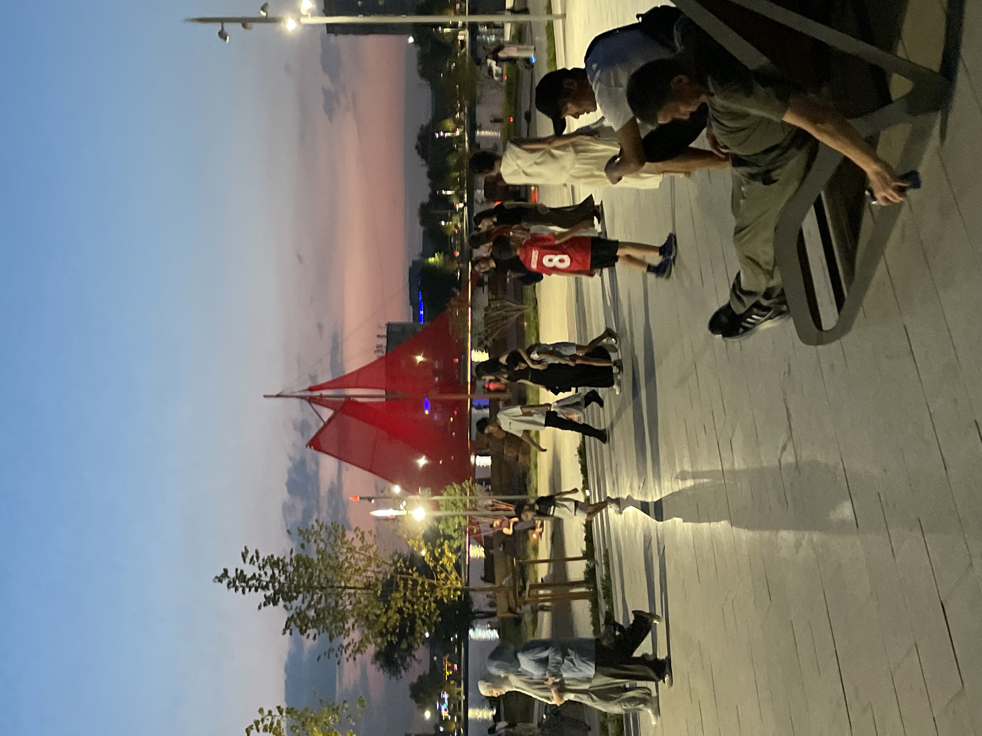 The image captures an outdoor scene at dusk, featuring a waterfront promenade with several people walking and relaxing. In the background, a large red sail-like structure is visible, possibly part of an art installation or architectural element, silhouetted against the twilight sky. The sky displays a gradient of blue to pink, indicating the transition from day to night. Lampposts are illuminated, casting light on the paved walkway.
Several individuals are present:
* Two women in the foreground are walking to the left, one wearing a light blue headscarf and the other a grey headscarf and loose grey clothing.
* A young boy in a red t-shirt with the number "8" on it is walking towards the right, accompanied by another child.
* Several other people, including adults and children, are dispersed throughout the promenade, some walking and others standing.
* In the immediate foreground, a man is reclined on a modern lounge chair, holding a dark object that appears to be a beverage can. He is looking downwards. Another person is partially visible next to him.
The environment includes a paved walkway with reflective surfaces, suggesting it might be wet or polished. Trees line the walkway and the waterfront. Beyond the promenade, a body of water reflects the lights from the city and the sky. Distant buildings and lights are visible across the water. The overall atmosphere is one of a public space being enjoyed during the early evening.
While the image depicts a scene in a city, the specific location in Almaty, Kazakhstan cannot be definitively determined from the visual information provided in the image itself.