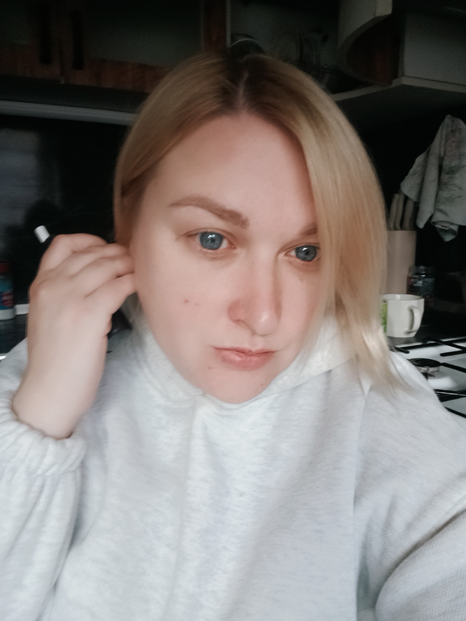 A woman with blonde hair and blue eyes is looking down at the camera, wearing a white hooded sweater. She is holding a white object near her ear with her right hand. Behind her is a kitchen setting with dark cabinets, a white mug, and a stovetop. It is not possible to verify if the location is indeed Chaikovsky, Russia.