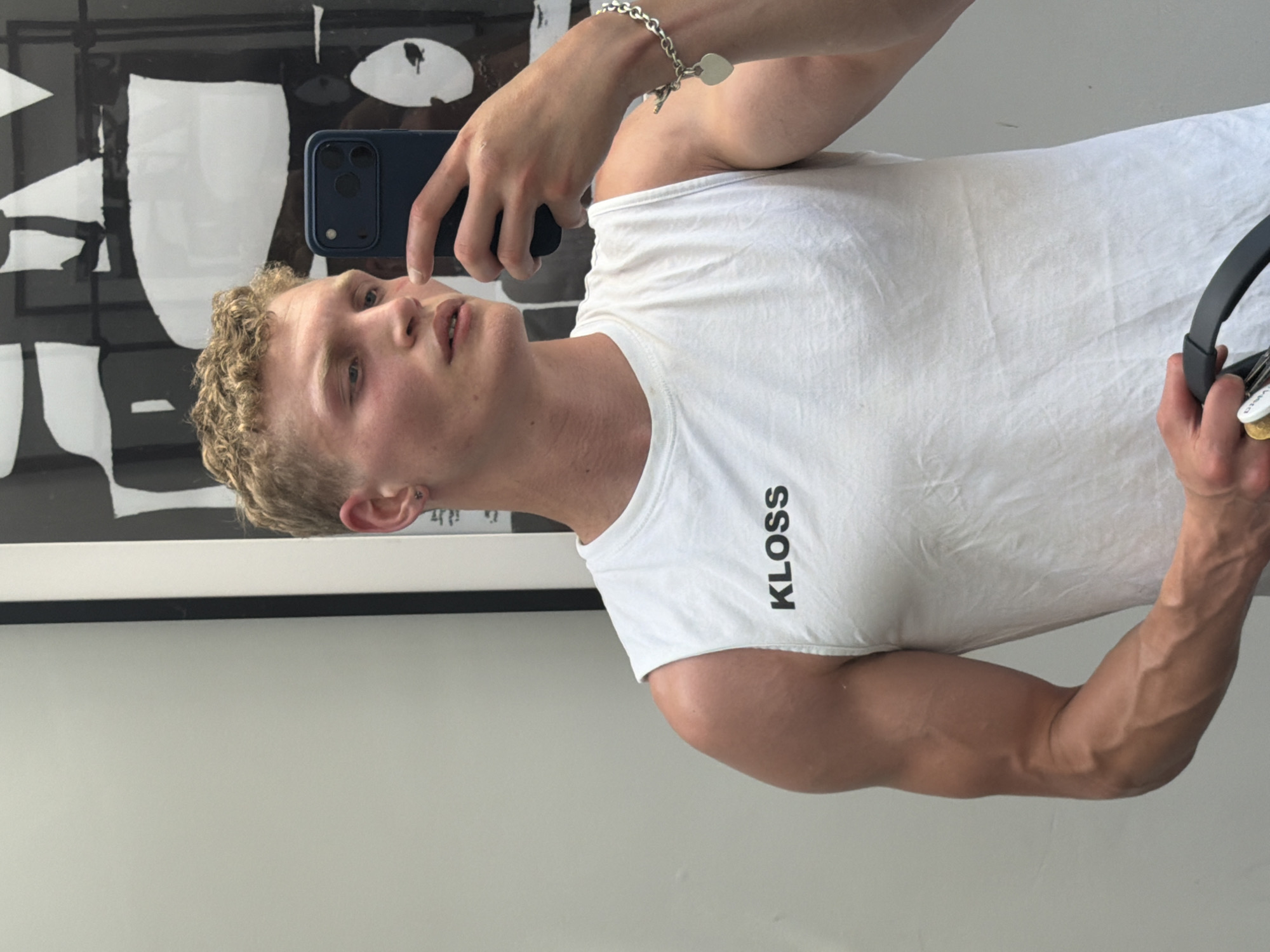 A fair-skinned person with short, curly blonde hair is taking a selfie indoors. The person is wearing a white sleeveless t-shirt with the word "KLOSS" printed vertically in black on the chest. The person is holding a dark blue smartphone in their left hand. On their left wrist, there is a silver bracelet with a heart-shaped charm. In the person's right hand, they are holding a pair of black and silver headphones. Behind the person, there is a black and white abstract art print framed in black. The background is a plain, light-colored wall. There is no information to determine the location as the city of London, United Kingdom.