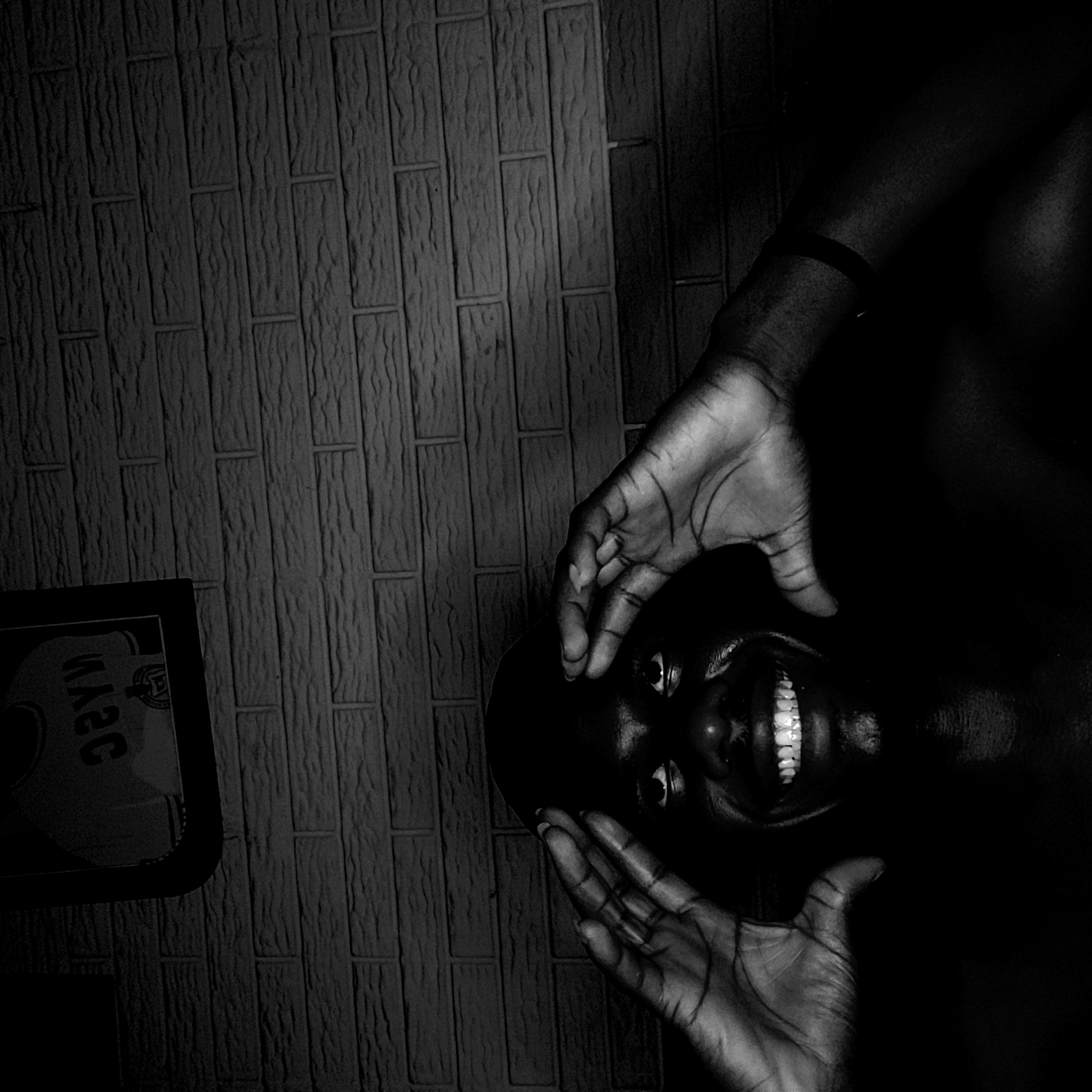 This monochrome, high-contrast portrait, captured in Narayi, Nigeria, features a person with dark skin. The person's head is tilted or upturned, presenting a wide, joyful smile that brightly illuminates their teeth. Both hands frame the face; one rests near the forehead or side of the head, while the other is positioned near the chin.
The lighting is dramatic, creating a stark chiaroscuro effect with intense highlights on the person's skin, hands, and teeth, contrasting sharply with the deep shadows that envelop much of the body and background. The backdrop is a textured wall, possibly constructed from bricks or blocks, displaying a distinctive vertical pattern that runs diagonally across the frame due to the image's rotation.
In the bottom-left corner, a reflective surface or object is visible, displaying reversed and partially legible text that appears similar to Cyrillic characters such as "УАЭС" or "СЭАУ". The overall impression conveyed by the image is one of happiness or playful energy, emphasized by the dramatic black and white aesthetic and the person's expressive facial features.