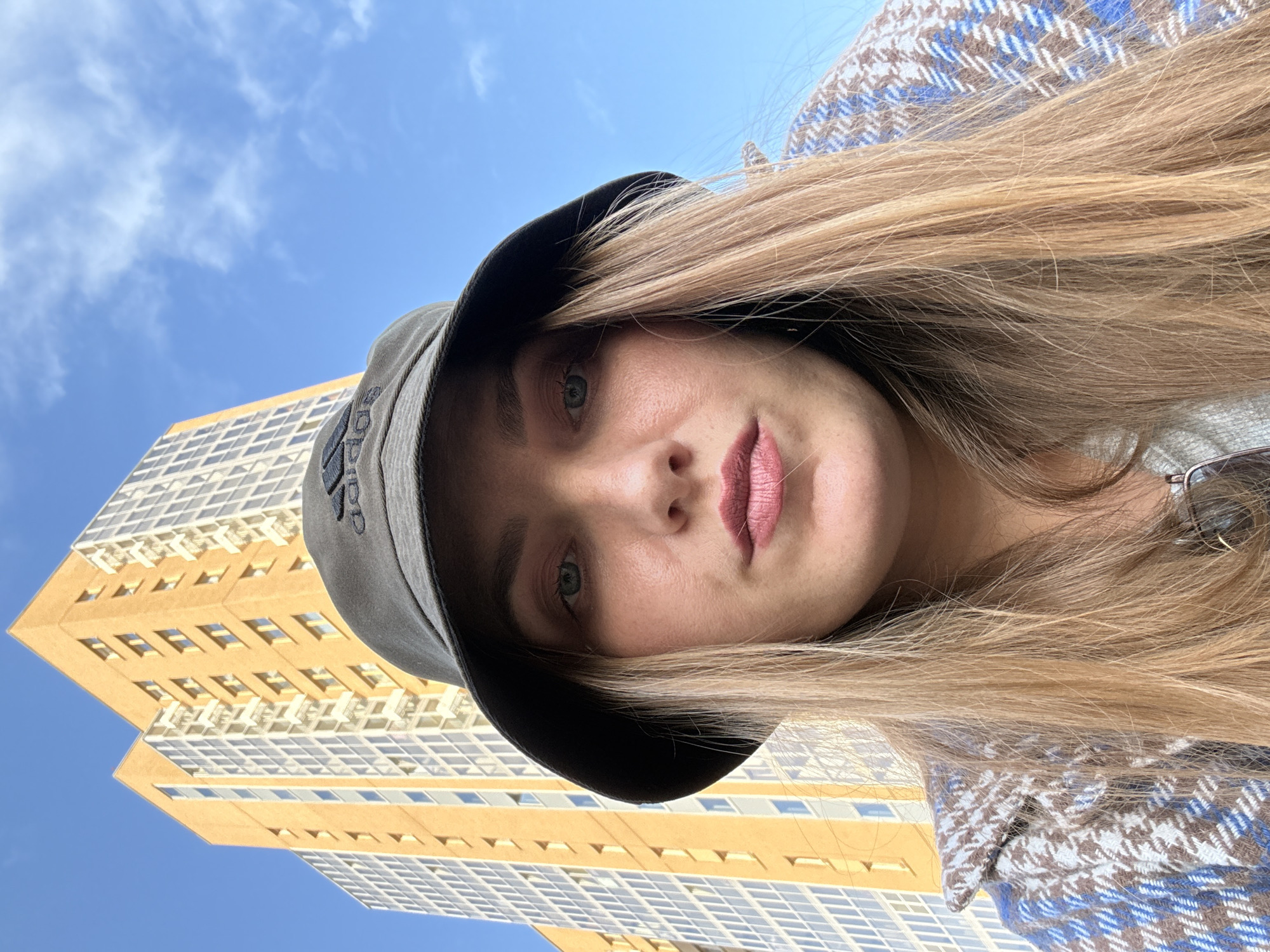 A young woman with blonde hair and wearing a dark bucket hat with an Adidas logo is looking directly at the camera. She has on a plaid jacket with blue, white, and brown patterns. Behind her, a tall, yellow apartment building with many windows dominates the frame against a bright blue sky with scattered white clouds. The building appears to be located in an urban environment, likely in Kazan, Russia, given the architectural style common in the city. The image is a close-up selfie, with the woman's face and upper body filling most of the foreground.