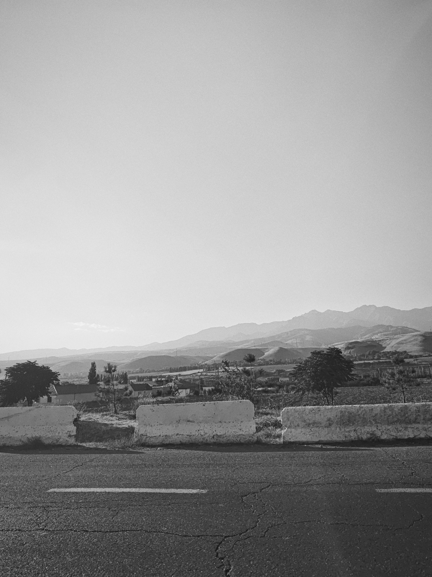 A black and white, medium-angle, outdoor shot captures a scenic view of the countryside from a roadside. In the foreground, a cracked asphalt road with a single white stripe runs horizontally across the bottom of the frame. A white concrete barrier, broken in the center, separates the road from the land beyond.
The middle ground reveals a rural landscape with a few scattered buildings and trees. Beyond that, a series of rolling hills and mountains recede into the distance, creating a layered effect. The sky is clear and bright, with a soft haze obscuring the farthest peaks. The image appears to be taken during daylight hours, likely in the late afternoon due to the soft lighting and long shadows. The overall atmosphere is serene and tranquil, evoking a sense of openness and natural beauty. The image does not contain any people or animals, nor any identifiable landmarks to pinpoint the exact location within Karankul, Uzbekistan.