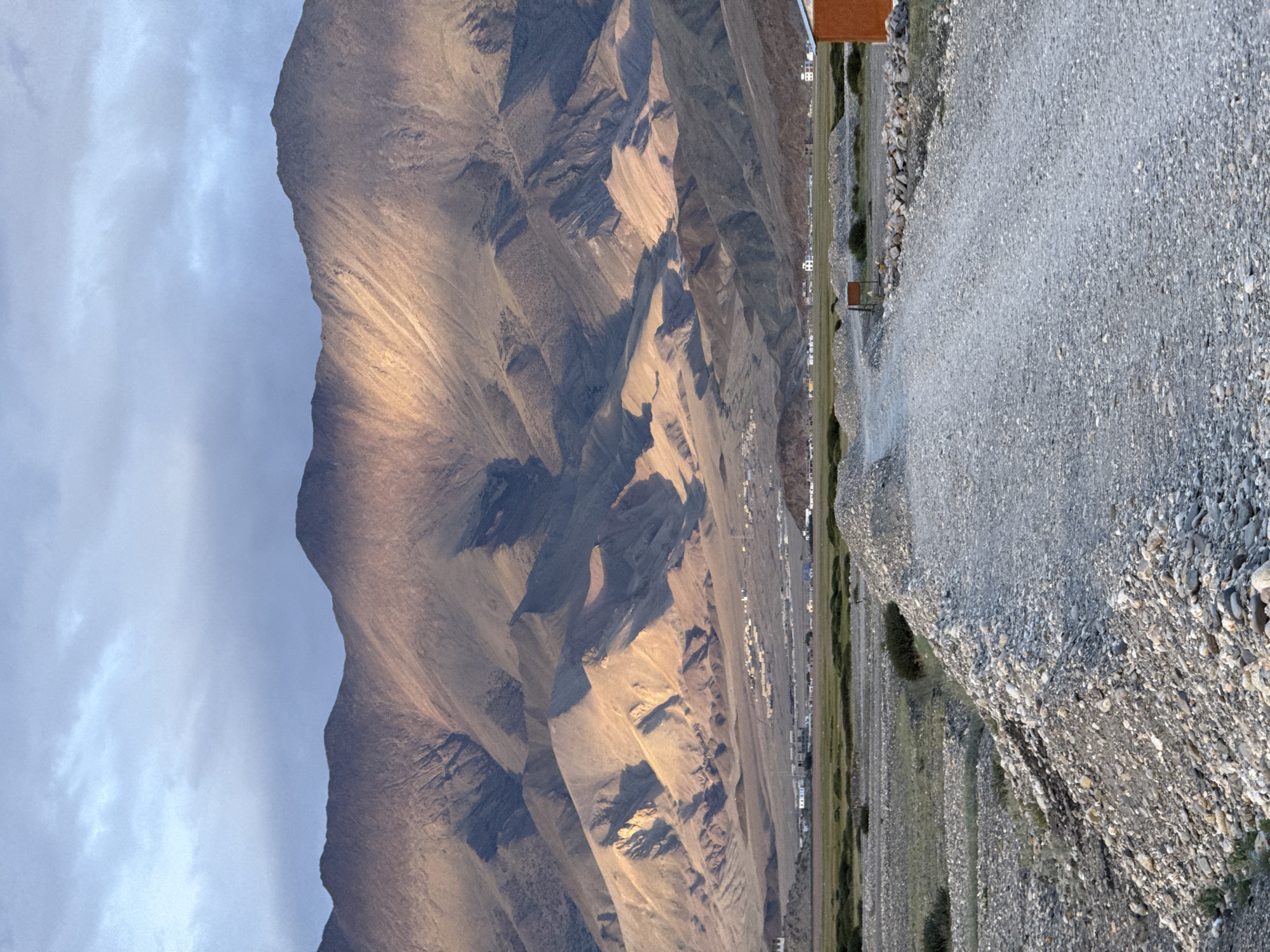 The image captures a vast mountainous landscape, likely near the city of Khuldo, India. In the foreground, a dirt road made of gravel leads towards the center of the frame, with piles of stones and sparse vegetation flanking it. The road appears to be an unpaved path through a semi-arid terrain.
In the middle ground, a flat, green valley stretches out, dotted with small clusters of buildings that suggest a village or settlement. The buildings are low-rise structures, some appearing to be made of stone or concrete. A few scattered trees or bushes break up the green expanse.
The background is dominated by a range of imposing mountains. The lower slopes of the mountains are rugged and uneven, showing signs of erosion and scree. The upper portions of the mountains are bathed in the warm, golden light of what appears to be either sunrise or sunset, creating dramatic shadows and highlighting the contours of the terrain. The sky above is partly cloudy, with soft grey and blue hues, indicating the time of day. The overall scene evokes a sense of remoteness and natural grandeur.