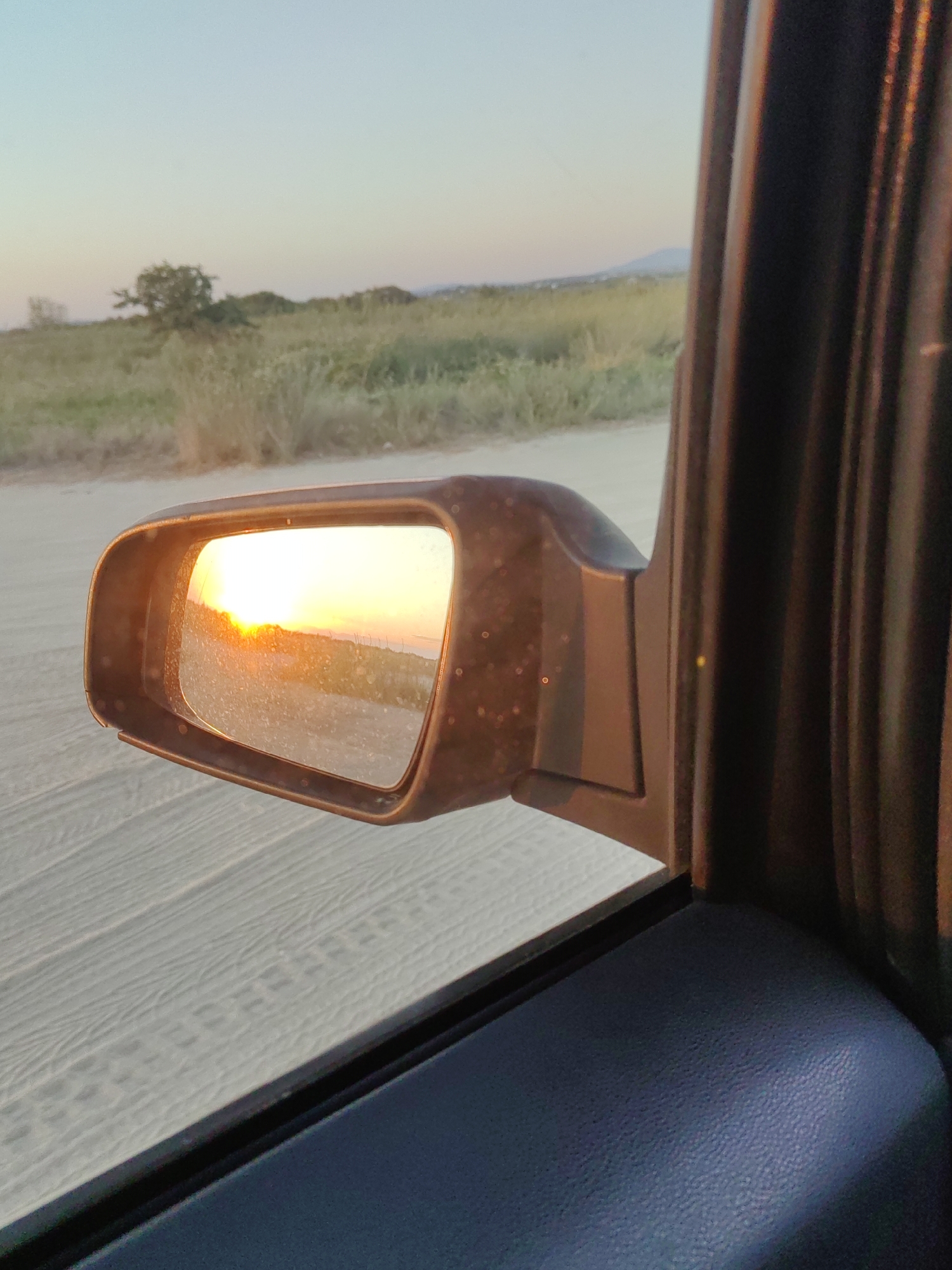 An outdoor, eye-level, medium shot captures the reflection of a sunset in a car's side mirror. The mirror is positioned on the right side of the frame, angled slightly towards the viewer. The reflection displays a horizon line with the sun low in the sky, casting an orange glow across a dusty landscape. The car's interior, including a dark gray door panel and part of a seat, is visible in the foreground and on the right edge of the image. The background outside the car, seen beyond the mirror and the car's window, shows a dry, grassy field with scattered bushes and trees. The ground outside the car appears to be a dirt road. The sky above the horizon is a soft gradient of pale blue and pink, indicative of twilight. The image was taken in Perea, Greece.