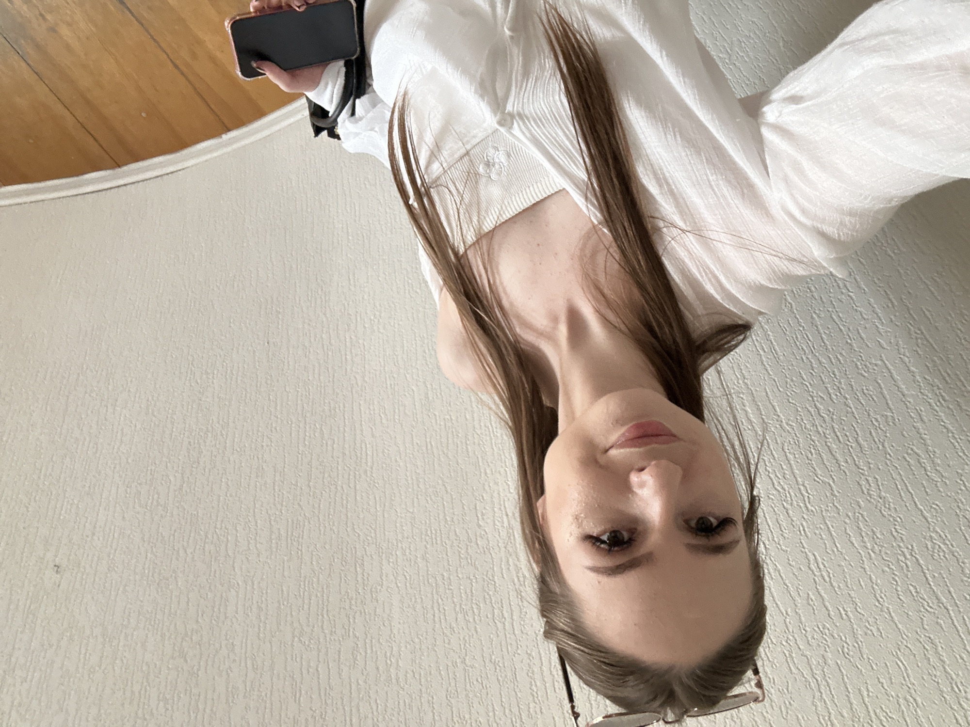 A young woman with long, brown hair is taking a selfie. She is wearing a white blouse and holding a black smartphone in her right hand. She is standing in front of a textured, off-white wall. The floor is made of light brown wood. A pair of glasses are resting on top of her head. The scene appears to be indoors.