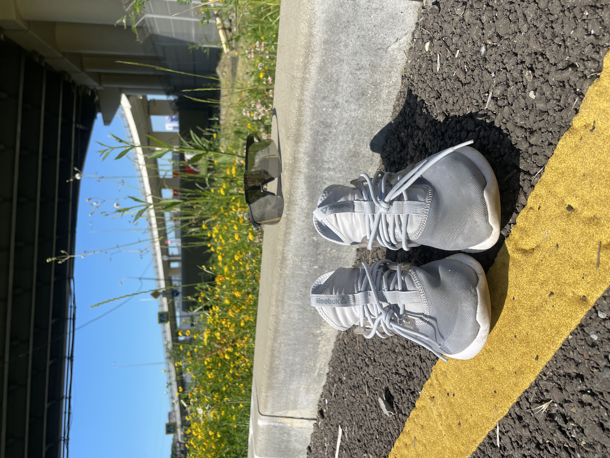 This rotated image captures a sunny, ground-level scene in Sochi, Russia, focusing on a pair of sneakers and their immediate surroundings.
In the foreground, a pair of light grey Reebok sneakers rests on a patch of dark asphalt, partially obscuring a bright yellow painted line that runs diagonally across the frame. The shoes appear well-worn but clean, with their laces tied. The brand name "Reebok" and its delta logo are clearly visible on the pull tab of the right shoe. Adjacent to the asphalt is a concrete curb, on which a pair of dark sunglasses lies. Beyond the curb, a vibrant strip of overgrown greenery with numerous small yellow flowers extends into the midground.
The background is dominated by a large, imposing concrete bridge or overpass structure, its architectural elements stretching across the frame under a clear, bright blue sky. The harsh shadows cast by the sneakers and the curb indicate strong, direct sunlight, suggesting a midday or early afternoon setting with clear weather. No people are visible, but the presence of the shoes and sunglasses implies a human presence, perhaps someone pausing during a walk or run. The scene conveys a sense of a quiet, sunny day in an urban outdoor environment, likely beneath or near a large piece of infrastructure in Sochi.