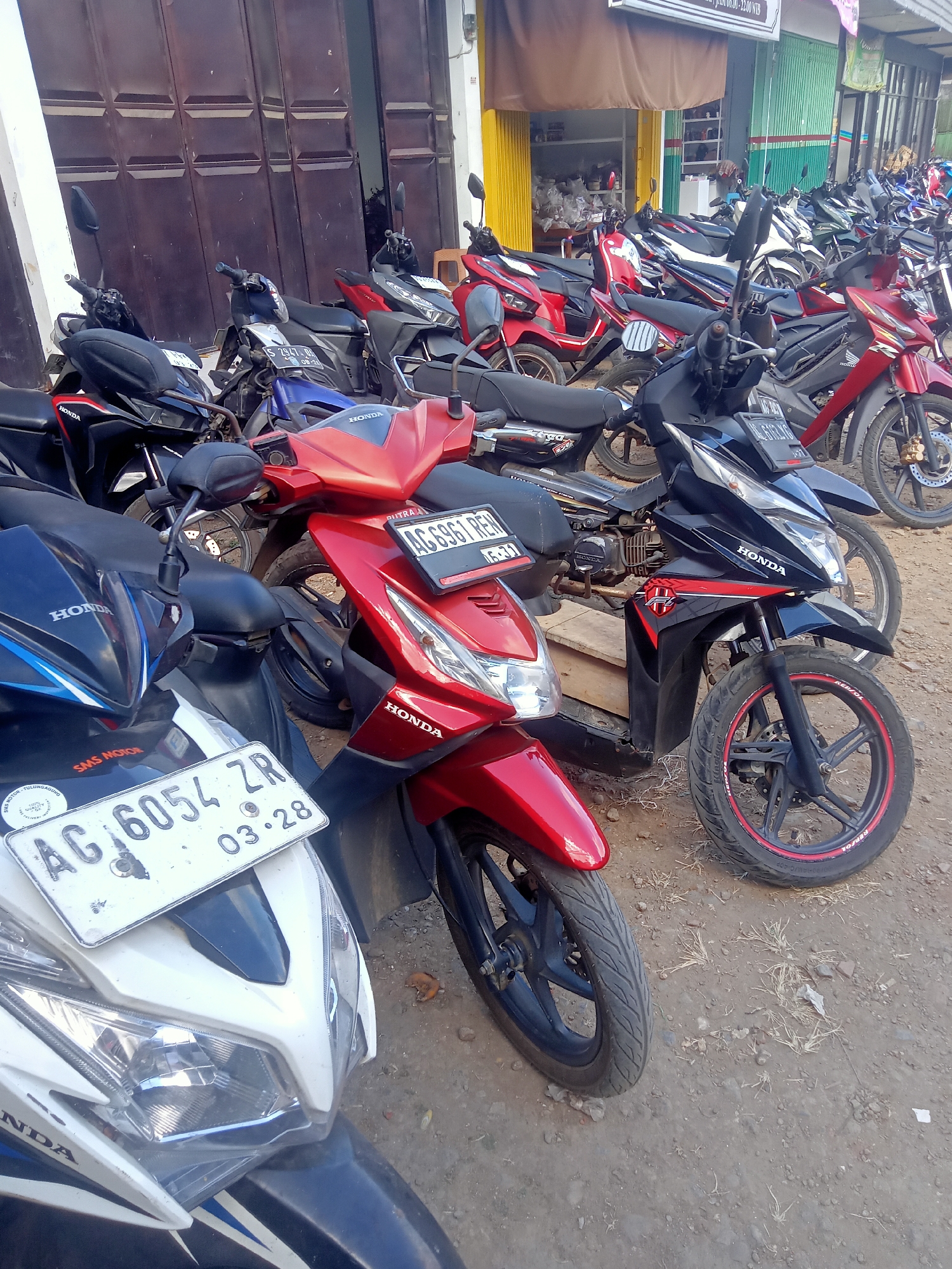 The image depicts a street scene in Pule, Indonesia, likely in front of a shop. Numerous motorcycles are parked in rows on a dirt and gravel surface. The motorcycles are of various colors, including red, black, and white, and appear to be mostly scooters and smaller motorbikes. Many of the vehicles display visible "HONDA" branding. One white motorcycle in the foreground has a license plate reading "AG 6054 ZR" and "03.28". Another red motorcycle has a license plate that is partially visible, appearing to read "AG 6961 RE...". Behind the motorcycles, a building with large brown doors and a storefront with green and yellow awnings is visible. The environment appears to be an outdoor setting with dry grass and debris scattered on the ground. No people are prominently visible in the image.
