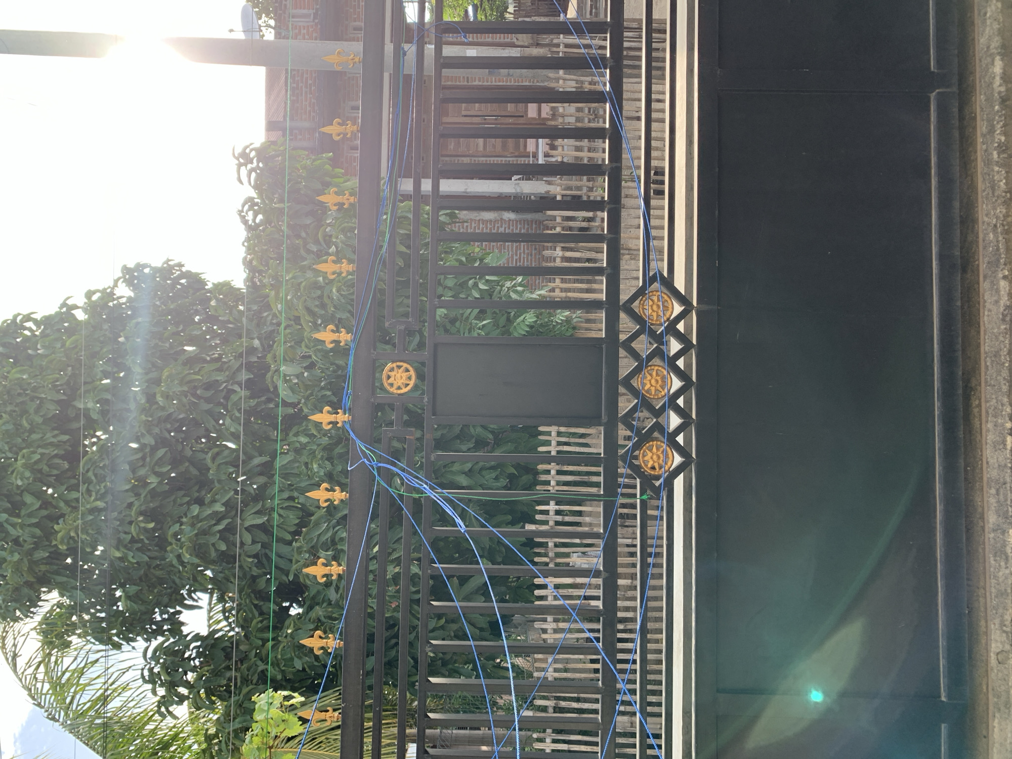 The image shows a black metal gate in front of a building, likely in Meunasahdrang, Indonesia. The gate is decorated with gold fleur-de-lis ornaments along the top and gold circular designs along the bottom. Several blue and green strings are stretched across the top of the gate, attached to the ornaments and other points. Behind the gate, a fence made of vertical wooden slats can be seen, along with the facade of a building constructed of red brick. Lush green trees and foliage are visible in the background, with a bright, hazy sky suggesting daylight. The image is shot from a slightly low angle, looking up at the gate and its surroundings.