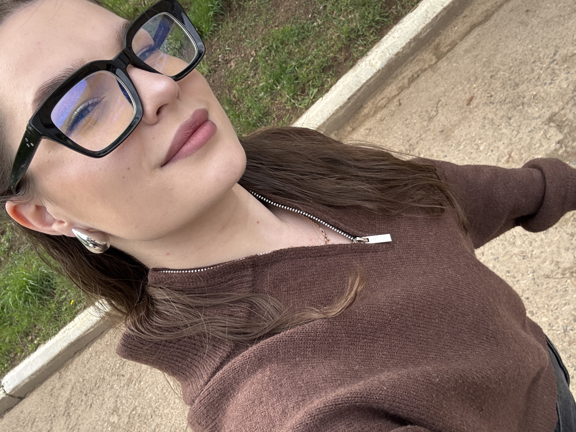 A young woman with fair skin and brown hair is depicted in a medium, angled selfie. She wears large, rectangular black-rimmed glasses. Her lips are painted with a mauve lipstick. She has a silver earring in her left ear.
The woman is wearing a brown zip-up sweater, with the zipper partially open, revealing a gold necklace.
The background suggests an outdoor setting, possibly a park or garden. There is a patch of green grass and a paved surface. The location is Chaikovsky, Russia.