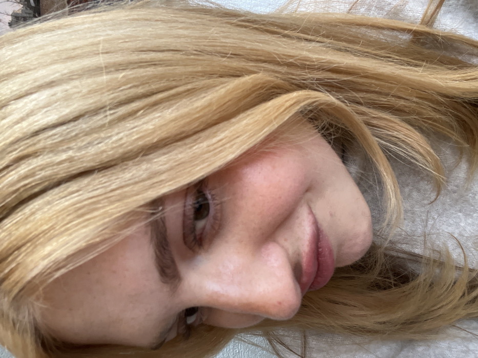 A close-up portrait shows a woman with blonde hair. Her face is tilted slightly to the side, revealing a profile view of her nose and lips. She has fair skin and light eyebrows. One eye is visible, displaying a brown iris. The blonde hair is voluminous, framing her face and cascading around her head. The background appears to be a soft, textured surface.