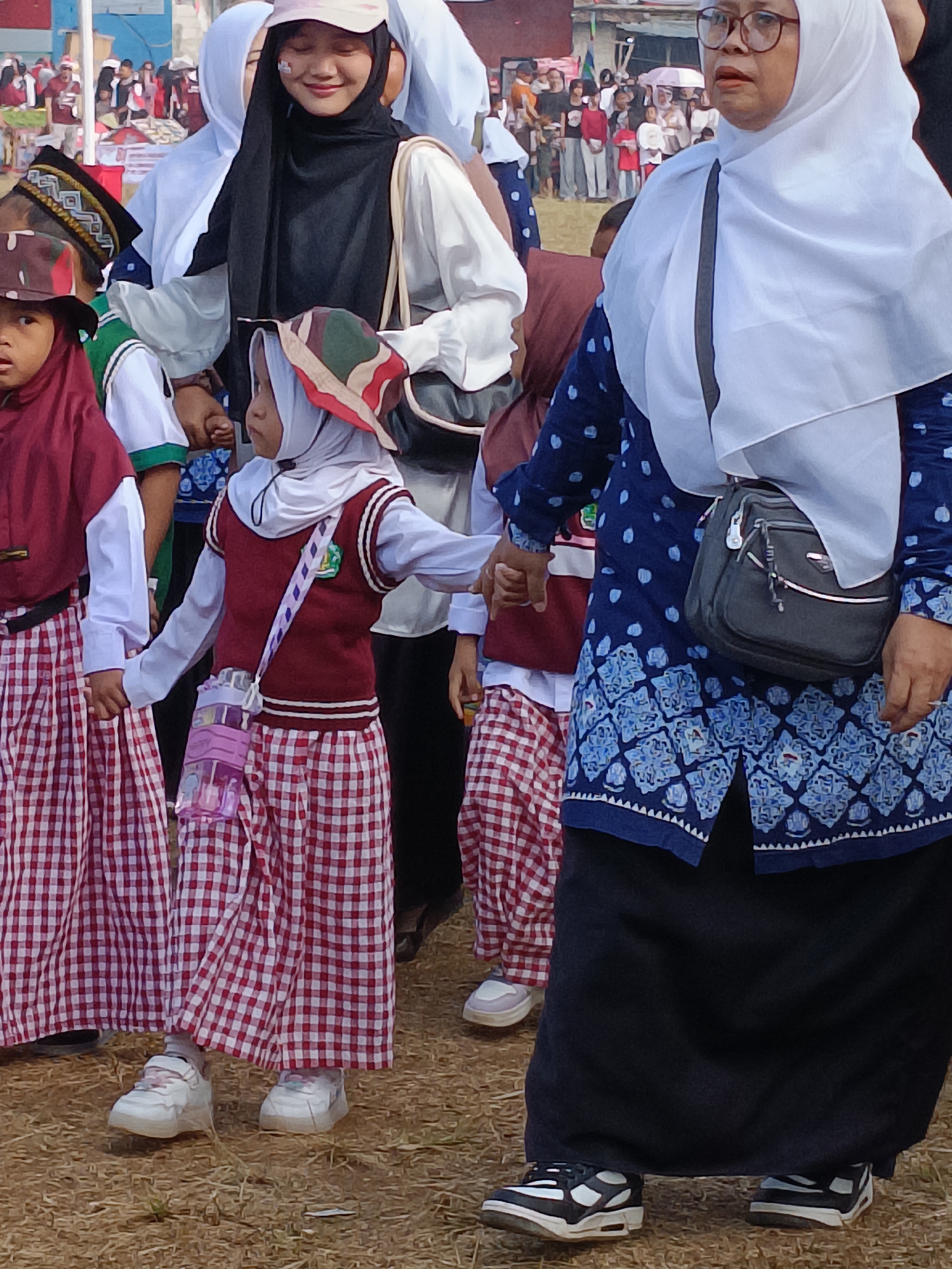 In Cibodas, Indonesia, a group of young girls wearing school uniforms and hijabs are participating in an outdoor event. Their uniforms consist of red and white checkered skirts and maroon vests over white long-sleeved shirts. One girl in the foreground holds hands with another girl beside her and carries a pink water bottle on a strap. They are walking on a grassy ground with dry leaves scattered around. Behind them, a woman in a blue patterned dress and white hijab, wearing glasses and carrying a black crossbody bag, is also present. Further in the background, more people and tents are visible, suggesting a community gathering or festival.