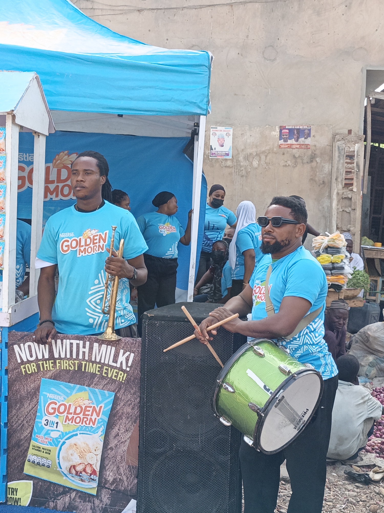 The image shows a promotional event for Nestle Golden Morn cereal in Kawaji, Nigeria. Two men in matching blue t-shirts with the Golden Morn logo are the focal point. The man on the left holds a trumpet while the man on the right is playing a green snare drum, positioned in front of a black speaker. A promotional stand with a "NOW WITH MILK!" poster is visible to the left of the men. Other people wearing similar blue t-shirts are visible behind the stand. In the background, there is a concrete wall with two posters. A crowded market scene is partially visible on the right, with piles of onions and people sitting or standing.