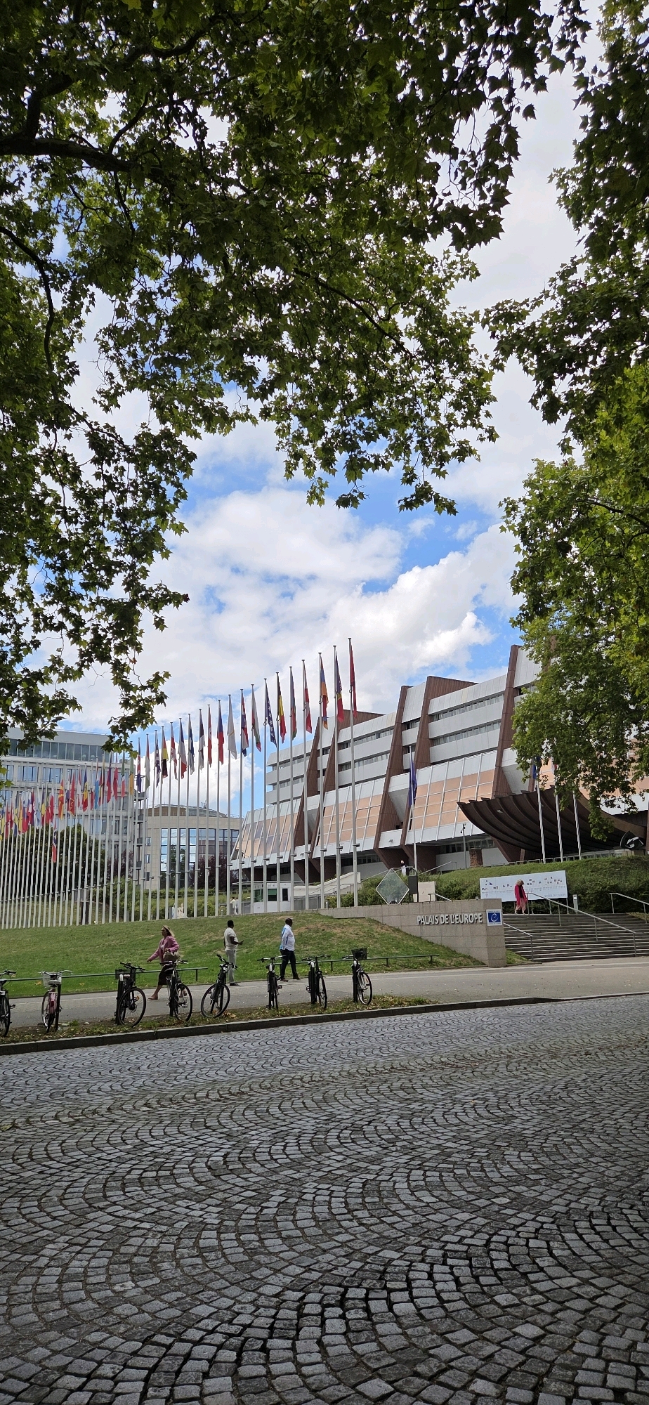 The image shows a wide view of the Palais de l'Europe in Strasbourg, France, a building adorned with flags from various European countries. The foreground features a cobblestone pathway leading to a paved road lined with bicycles. In the middle ground, a grassy area separates the road from the building, with several people walking and standing. The Palais de l'Europe itself is a large, modern building with a distinctive architectural style, featuring angled facades and numerous windows. The sky above is partly cloudy with blue patches. The overall scene conveys a sense of an institutional setting with a public, accessible area.