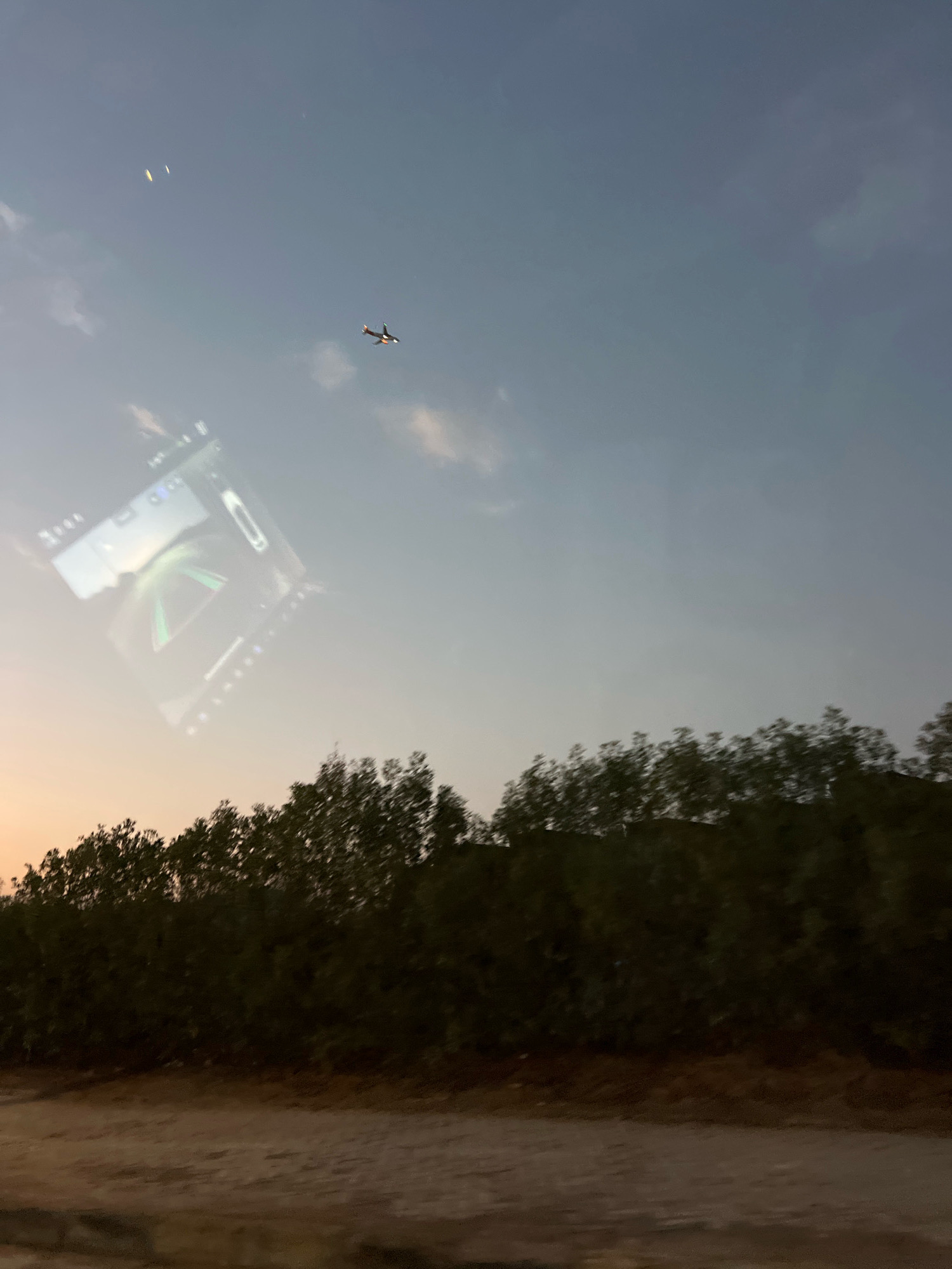 The image depicts a serene sky at dusk, with a small airplane in flight and a reflection of a digital display, possibly a car's infotainment system, superimposed on the sky. A dense line of trees borders the bottom of the frame, with a patch of sandy ground in the foreground. The scene is likely taken from within a vehicle, given the reflection. The lighting suggests an outdoor setting, perhaps on the outskirts of Sharm El Sheikh, Egypt, where the natural beauty of the sky and landscape is a prominent feature.