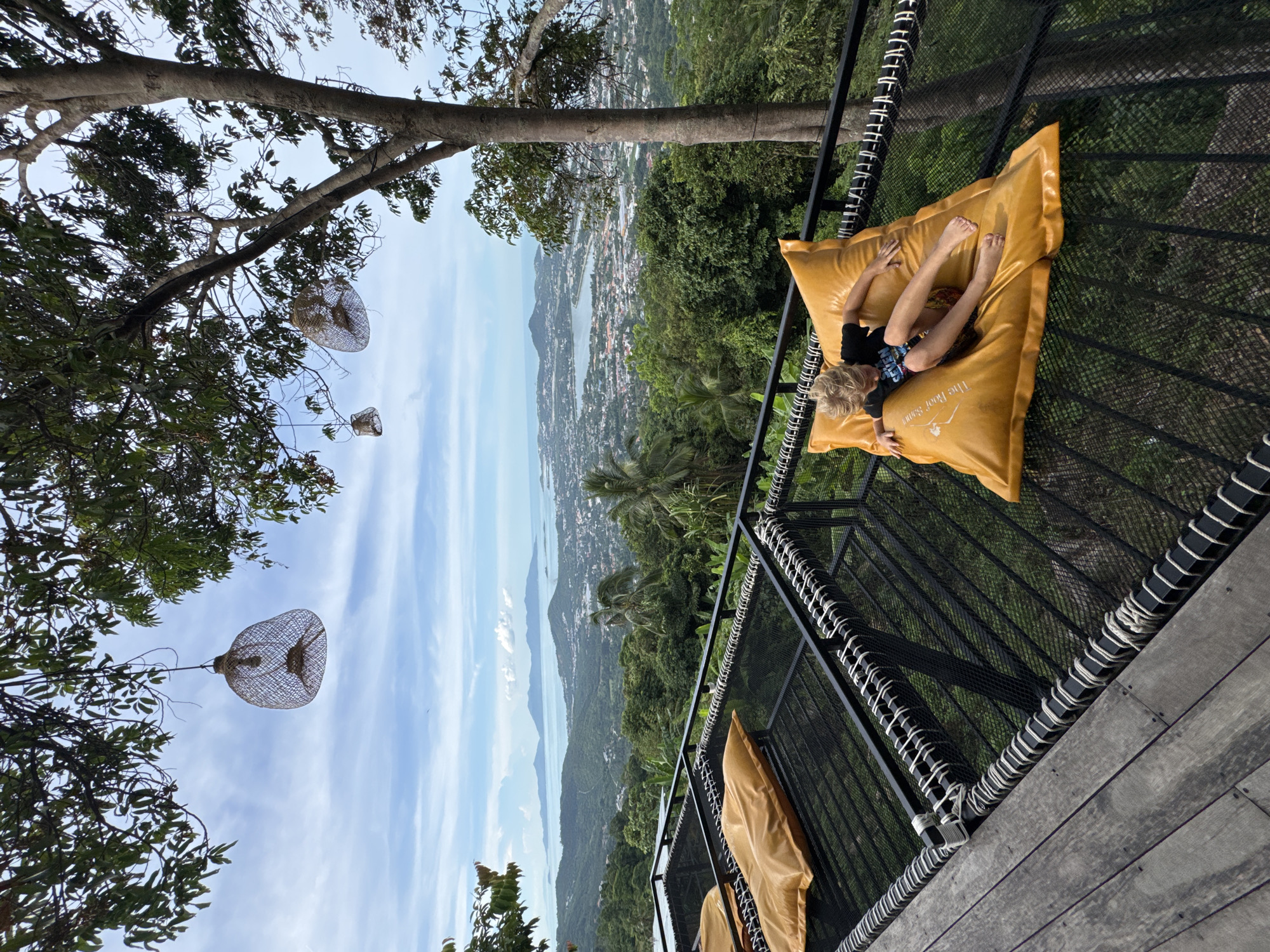An outdoor scene captures a young blonde boy lying on a large, orange beanbag on a netted platform overlooking a sprawling city nestled among lush green hills and a wide expanse of the ocean. The boy is wearing a dark t-shirt and patterned shorts. In the foreground, the wooden floorboards of the platform are visible, along with more orange beanbags. The background reveals a hazy cityscape dotted with buildings, leading to the blue ocean with distant islands under a cloudy sky. Overhead, tree branches with green leaves extend into the frame, and woven bamboo light fixtures hang from the branches.