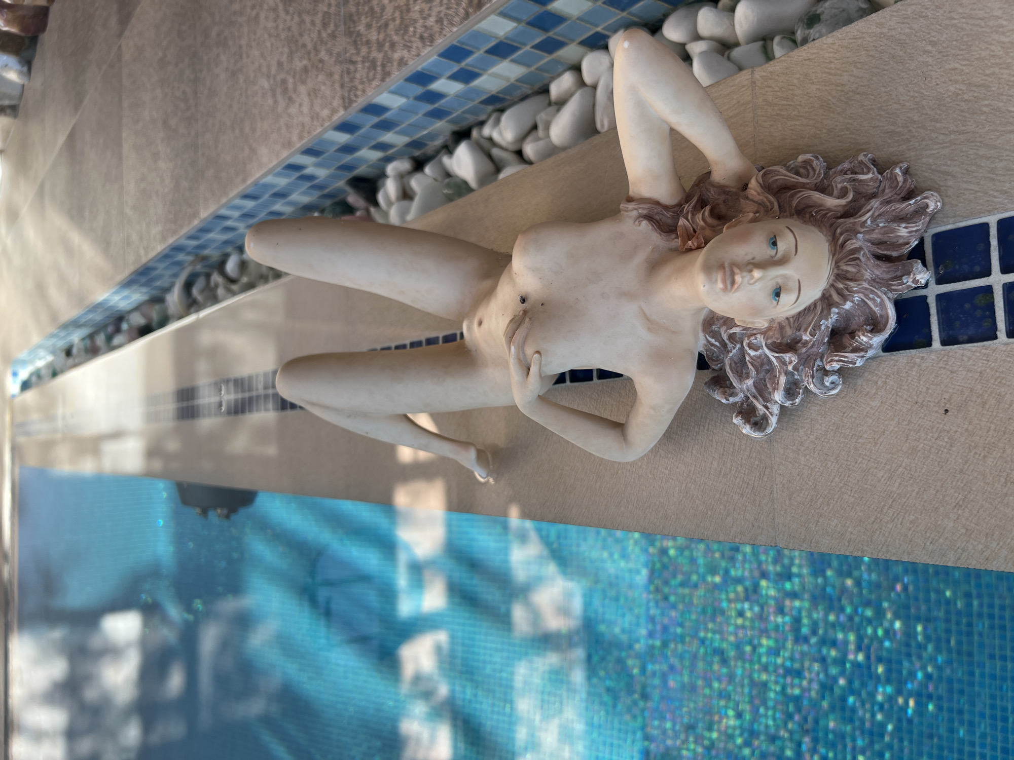 A nude statue of a woman is lying on the edge of a swimming pool, with her legs extended towards the water. The statue has light-colored skin, blue eyes, and long, wavy brown hair. Her right arm is bent, with her hand resting on her chest, and her left arm is bent behind her head. The pool is filled with blue water, and sunlight reflects off the surface. The edge of the pool is tiled with blue and white squares, and smooth white stones are visible in a narrow trough next to the statue. The scene appears to be outdoors, likely in a private backyard or resort in Bender, Moldova.