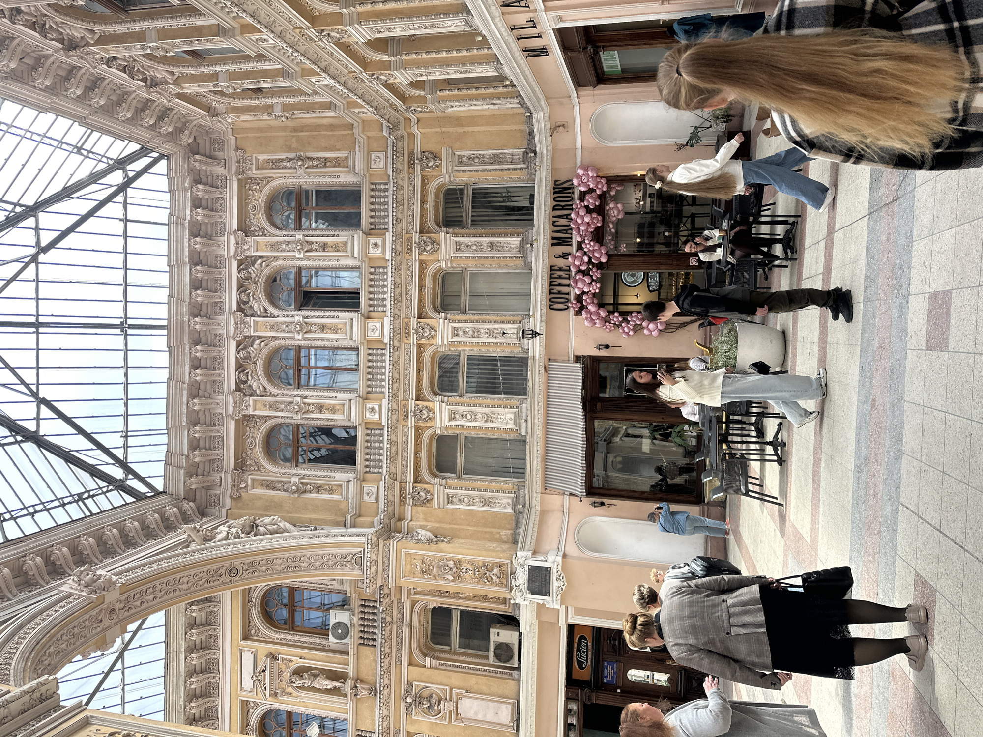 The image captures a bustling scene inside the Passage in Odesa, Ukraine. The ornate architecture of the passage is visible with intricate carvings and a glass ceiling allowing natural light to illuminate the area. Several people are visible, including individuals standing and walking through the passage, and some seated at tables outside a cafe or shop called "Coffee & Macarons." A woman with long blonde hair and a person wearing a plaid coat are in the foreground. Another store named "Lucien" is visible further down the passageway. Decorative pink balloons adorn the front of the cafe.