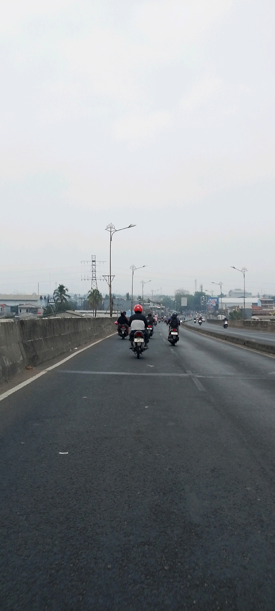 A group of motorcyclists are traveling on a highway in Bekasi, Indonesia. The highway has multiple lanes and is bordered by a concrete barrier on the left. The sky is overcast and the overall lighting is dim, suggesting it may be early morning or late afternoon. In the background, there are industrial buildings, power lines, and utility poles, characteristic of an urban or semi-urban industrial area in Bekasi. The motorcyclists are wearing helmets and appear to be traveling in the same direction. The scene is captured from a low-angle perspective, likely from another motorcycle.