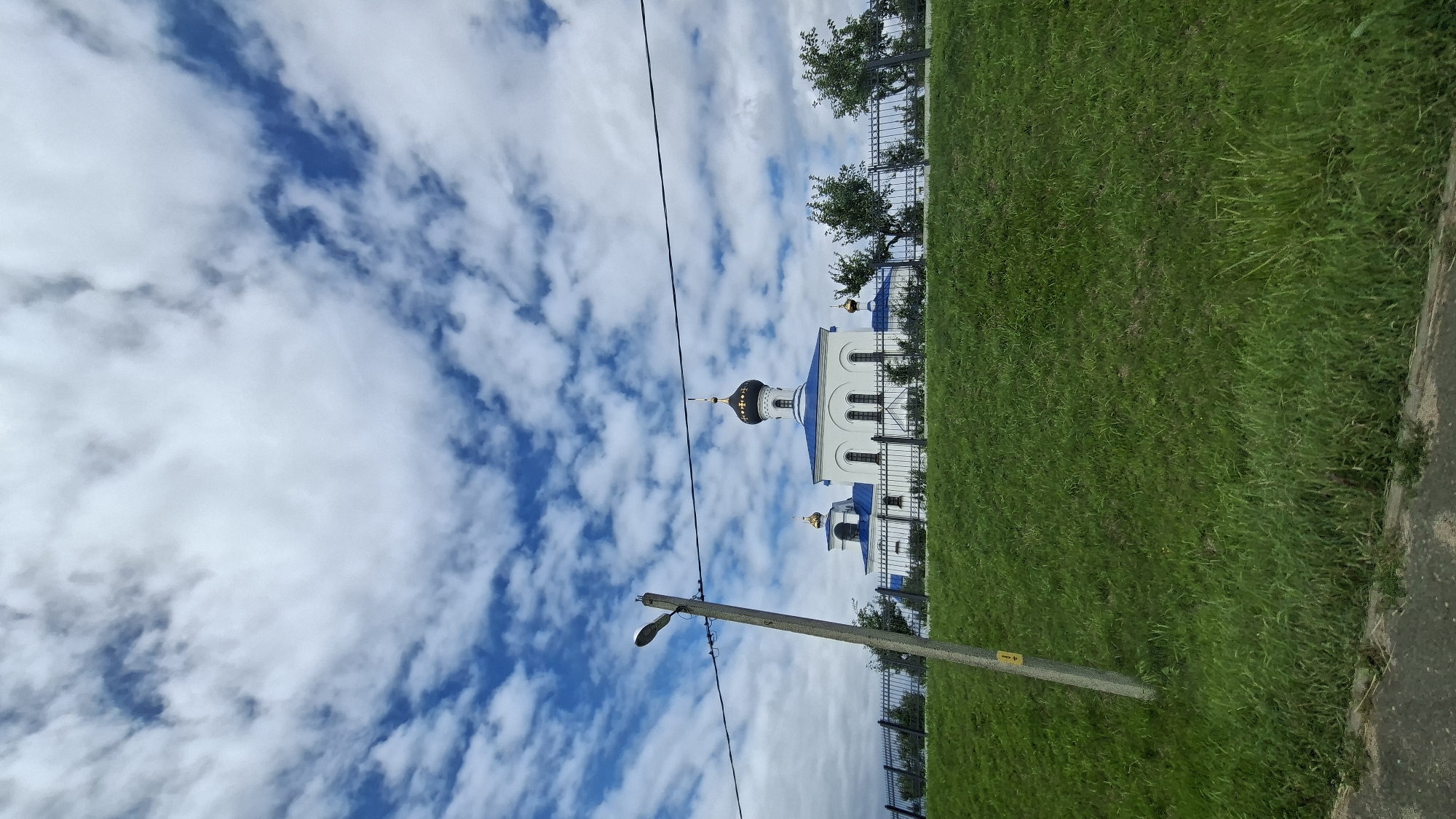 The image depicts a white church with a blue roof and golden domes, located on a grassy hill behind a black metal fence. The sky above is blue with white, scattered clouds. In the foreground, a gray concrete utility pole with a lamppost stands to the left of the frame, with a power line running horizontally across the image. Lush green grass covers the sloping terrain in front of the fence and the church. The environment suggests an outdoor setting, possibly on the outskirts of a town or in a park-like area. The architectural style of the church is characteristic of Eastern Orthodox churches. The scene appears to be tranquil and scenic.