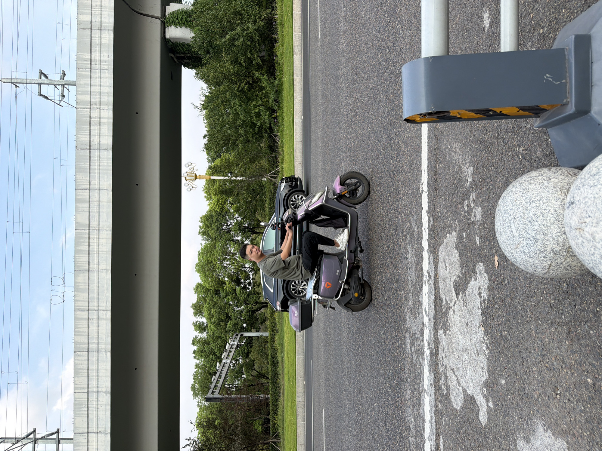 A young adult male rides a purple electric scooter on a paved road in Jiangjin, China. He wears a dark olive t-shirt and black pants with white sneakers. He is looking towards the camera. Behind him, a black SUV is parked on the right side of the road. In the background, a concrete overpass supported by pillars spans the road. Trees and vegetation line the roadside, and overhead power lines are visible. The road surface is asphalt with white lane markings. In the foreground, a grey protective barrier with yellow reflective tape and two large speckled spheres are present.