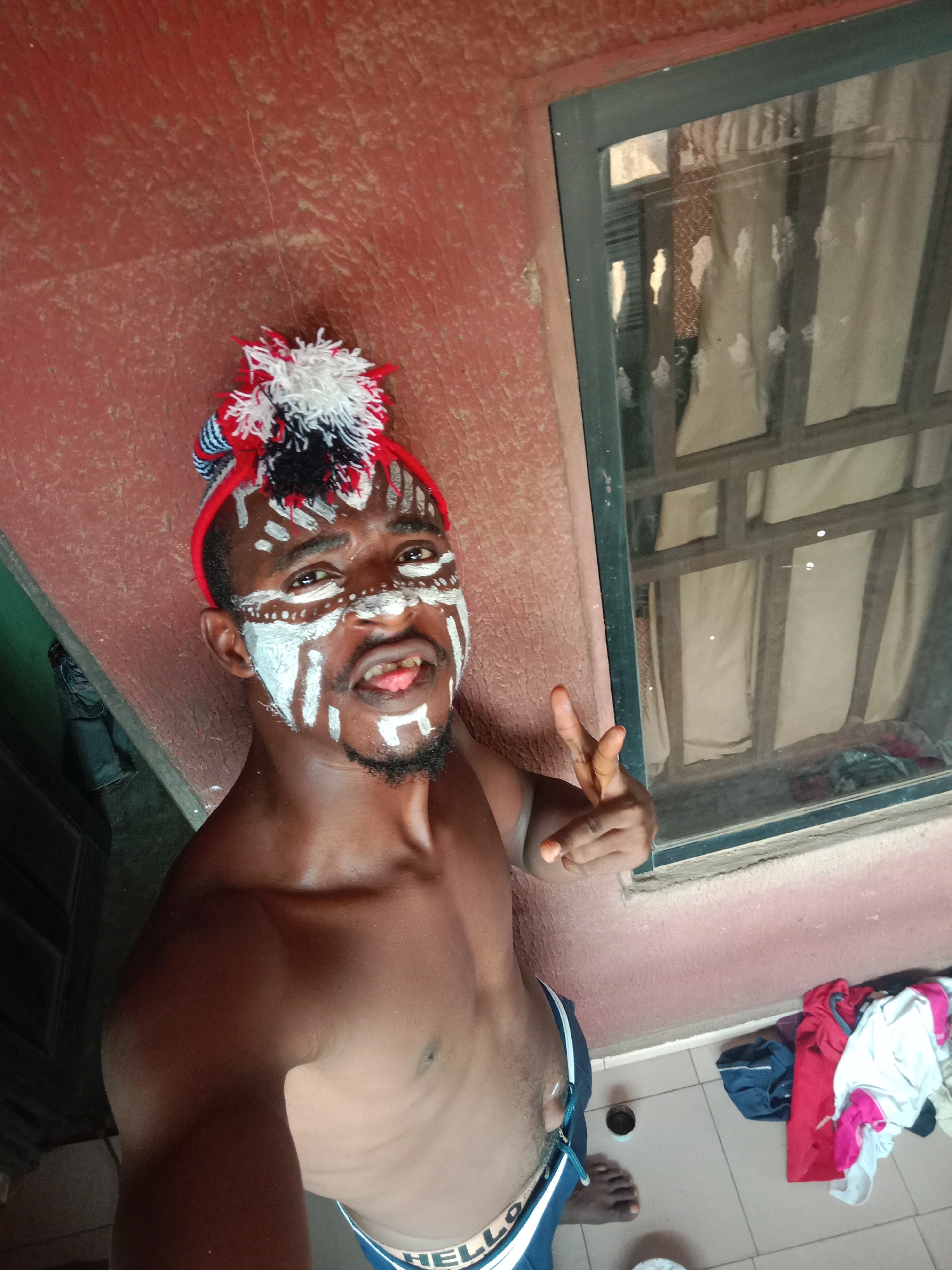 A shirtless, dark-skinned man with a black beard and white face paint is positioned in front of a reddish-brown wall and a window. The face paint consists of white stripes and dots around his eyes and mouth. He is wearing a red headband with a white and black pom-pom attached. He is sticking his tongue out and making a peace sign with his right hand. The window has a dark frame and a sheer curtain behind it. On the floor, to the right of the man, is a pile of clothing, including a red shirt, a white shirt, and a dark blue bag. The location appears to be indoors, as there is a tiled floor visible. No specific details about the location in the city of Unknown, Unknown, are evident.