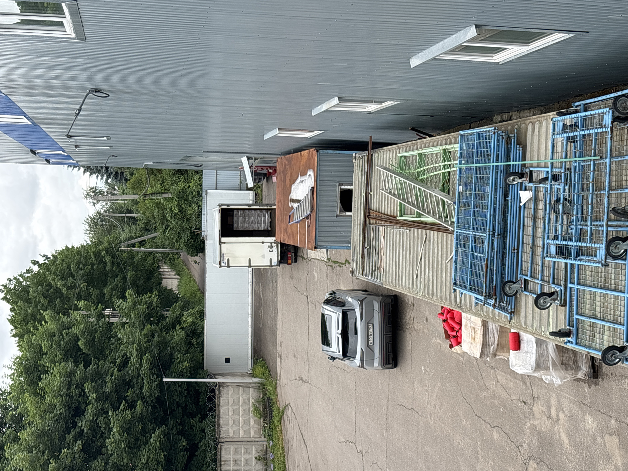 An aerial view captures a large, grey, corrugated metal industrial building with blue trim along the top. Several white-framed windows are visible on its side. In front of the building, a paved lot contains a modern grey SUV with a sunroof and a license plate reading "K 101EE 977". Adjacent to the SUV is a white truck with its back doors open, revealing stacks of boxed items. Further into the lot, a small, rusty-brown shed-like structure with a damaged roof sits next to a larger white, corrugated metal structure. A concrete fence with barbed wire topping is visible in the background, with lush green trees and foliage behind it. Piled on a pallet near the front of the image are several large red conical objects, and stacked metal cages with blue frames and wheels are positioned in the foreground, resting on a grey, corrugated platform. The scene appears to be an industrial yard. The location is identified as Ostrov, Russia.