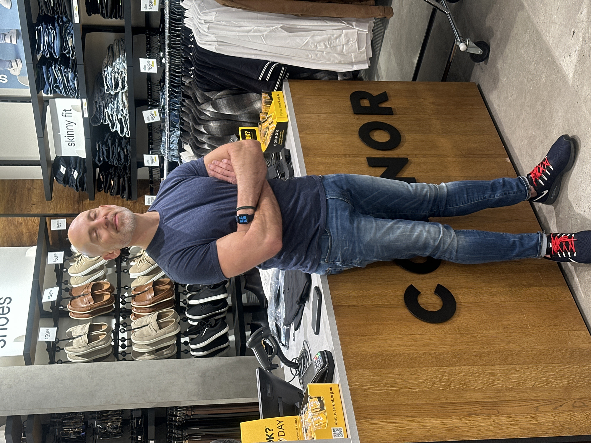 A bald, middle-aged Caucasian man with a beard stands with his arms crossed in front of a wooden counter inside a retail store. He is wearing a navy blue t-shirt and blue jeans, with dark athletic shoes with red laces. The store appears to be a men's clothing store, with racks of shirts and shelves of jeans and shoes visible in the background. The counter has a computer monitor, a barcode scanner, and a payment terminal on it. A yellow box with the words "R U OK?" is also on the counter. The store's name, "CONNOR," is visible in large black letters on the front of the counter. The man is looking slightly upwards with his eyes closed, a faint smile on his face. The store is clean and well-lit. The specific location in Coffs Harbour, Australia is not determinable from the image.