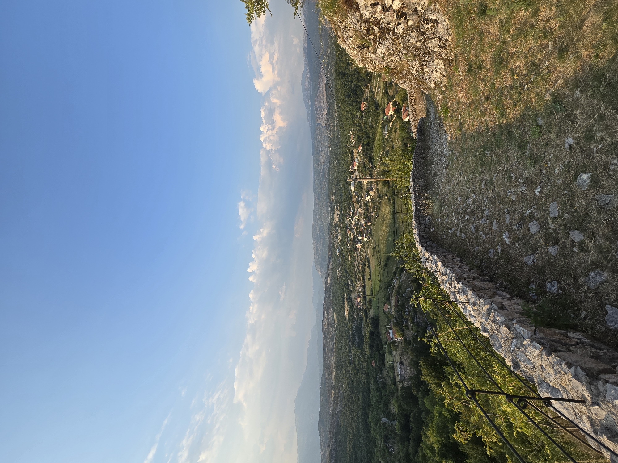 The image captures a panoramic view of a rural landscape, likely from an elevated vantage point in Gornja Vrbica, Montenegro. The scene is dominated by a vast expanse of rolling hills covered in dense green vegetation, interspersed with small clusters of red-roofed houses forming a village. The sky above is a clear, bright blue with scattered white, puffy clouds, suggesting fair weather. In the foreground, a stone wall and a paved pathway are visible, hinting at a constructed viewpoint or historical site. The pathway is uneven and appears to be made of cobblestones and dry grass. A black metal railing runs along the edge of the wall. In the distance, hazy mountains form the horizon. The overall impression is one of tranquility and natural beauty, characteristic of the Montenegrin countryside. No people or specific actions are depicted in the image.