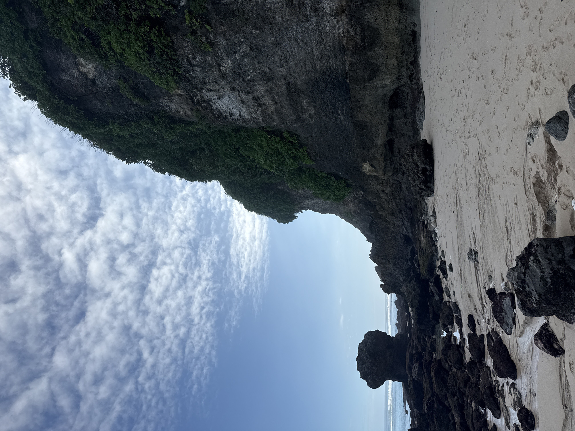 The image shows a view of a beach from under a rocky overhang in Kuta Selatan, Indonesia. The overhang is covered in green vegetation. The beach is sandy with scattered dark rocks. The sky is blue with scattered white clouds. The ocean is visible in the distance. The image is taken from a low angle, looking upwards and outwards from the overhang.
