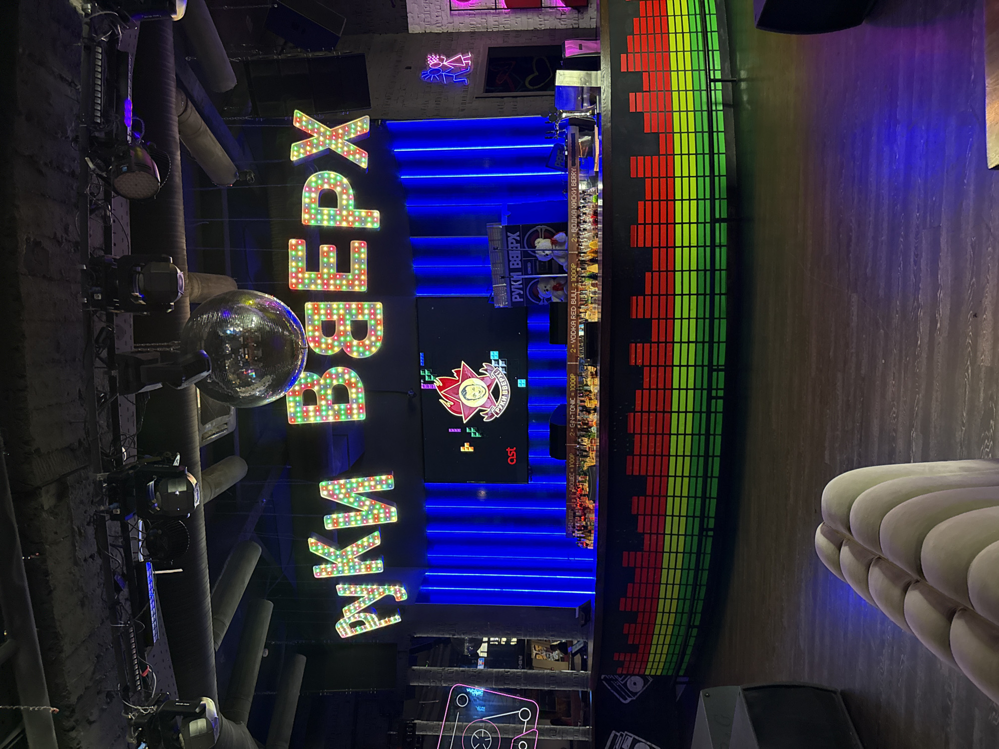 The image shows the interior of a bar or nightclub in Moscow, Russia, judging from the Cyrillic lettering on the signage. The dominant feature is the large, illuminated sign "РУКИ ВВЕРХ" (Ruki Vverkh), indicating it is likely themed around the Russian pop group of the same name. The sign is lit with colorful bulbs and sits above a bar area illuminated with blue neon lights. Behind the bar, there is a screen displaying a stylized image related to the "Ruki Vverkh" theme, along with drink advertisements in Cyrillic. There is also a disco ball and lighting fixtures hanging from the ceiling. Adjacent to the bar is a large, colorful display resembling a graphic equalizer with red, green, and yellow bars. A rounded booth seating area is visible on the right side of the image. The overall atmosphere appears to be that of a lively, themed entertainment venue.