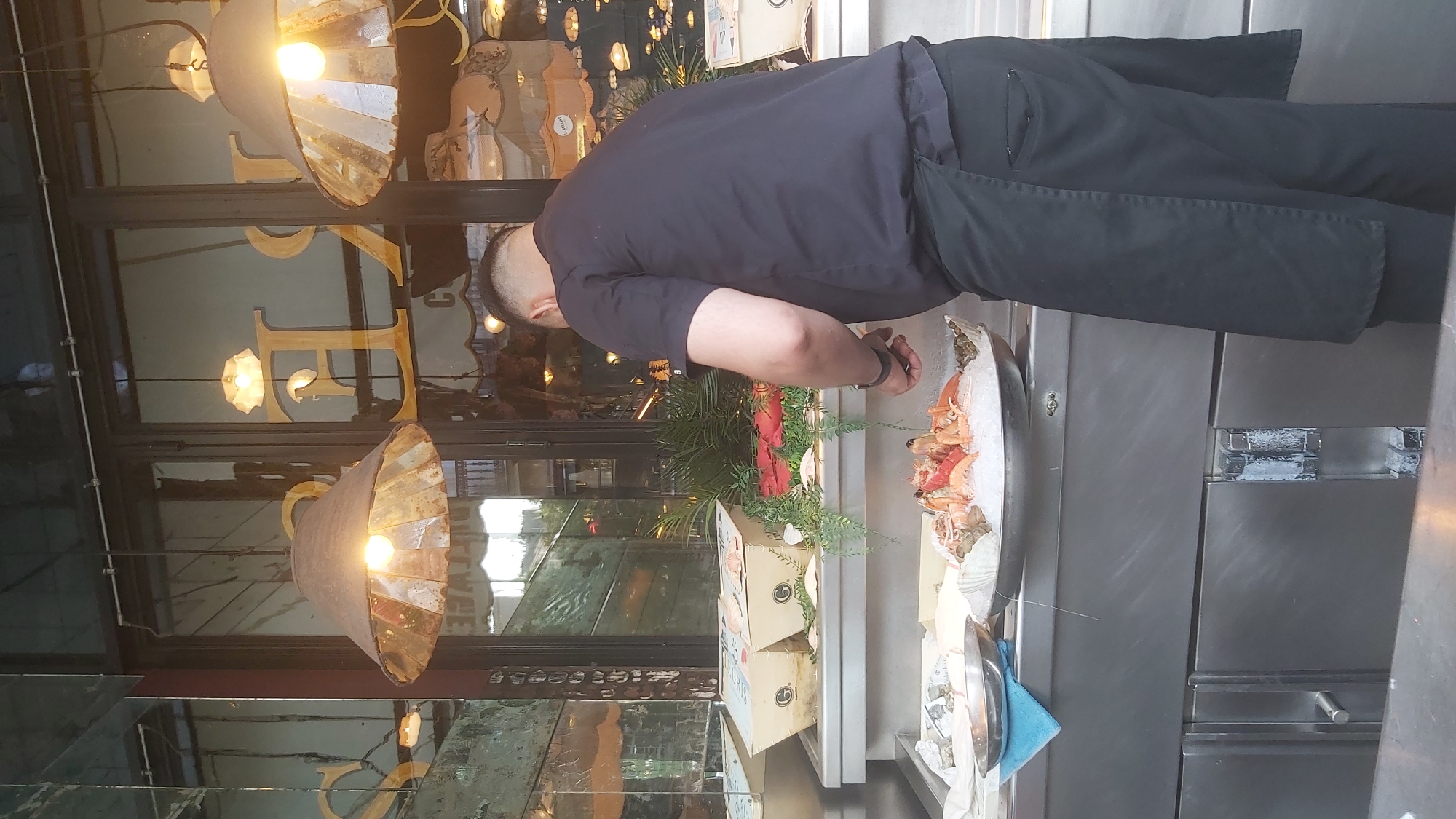 A male worker wearing a dark blue short-sleeved chef's coat and a black apron is standing in front of a display of seafood on ice. He is holding a knife and appears to be preparing the seafood. In the background, there is a glass storefront with large, ornate gold lettering that reads "LE". The storefront has reflections of lamps and interior details. The seafood display includes lobsters, prawns, and shellfish on a bed of crushed ice in a large silver platter. There are also cardboard boxes with seafood in them on a shelf above the display. The overall scene suggests an outdoor seafood market or restaurant in Paris, France.