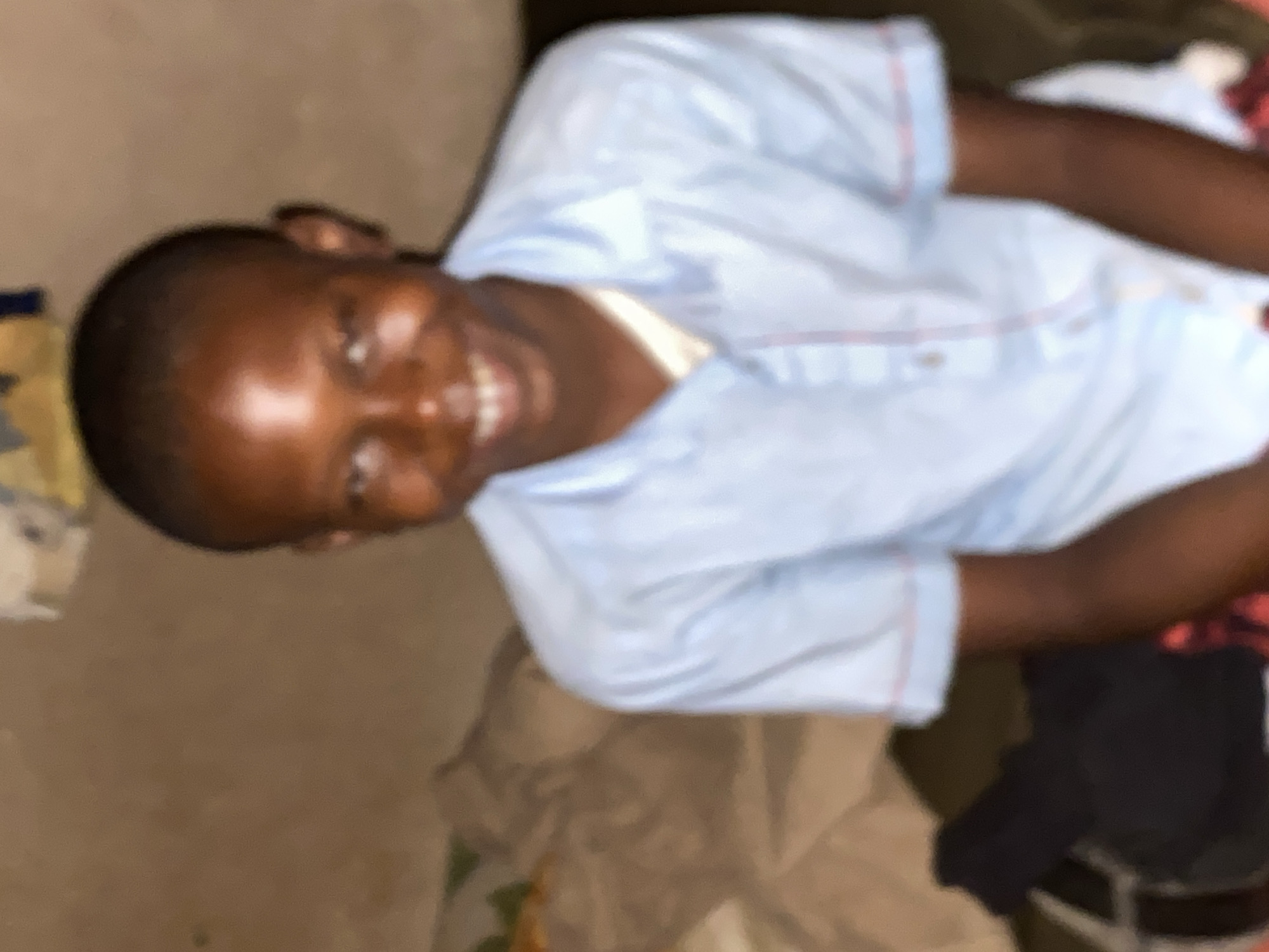 A young, dark-skinned person is lying down and smiling. The person is wearing a light blue button-down shirt with red trim. The person is surrounded by miscellaneous items including clothing and possibly bedding. The location is stated to be Bukuru, Nigeria. No further information is discernible.