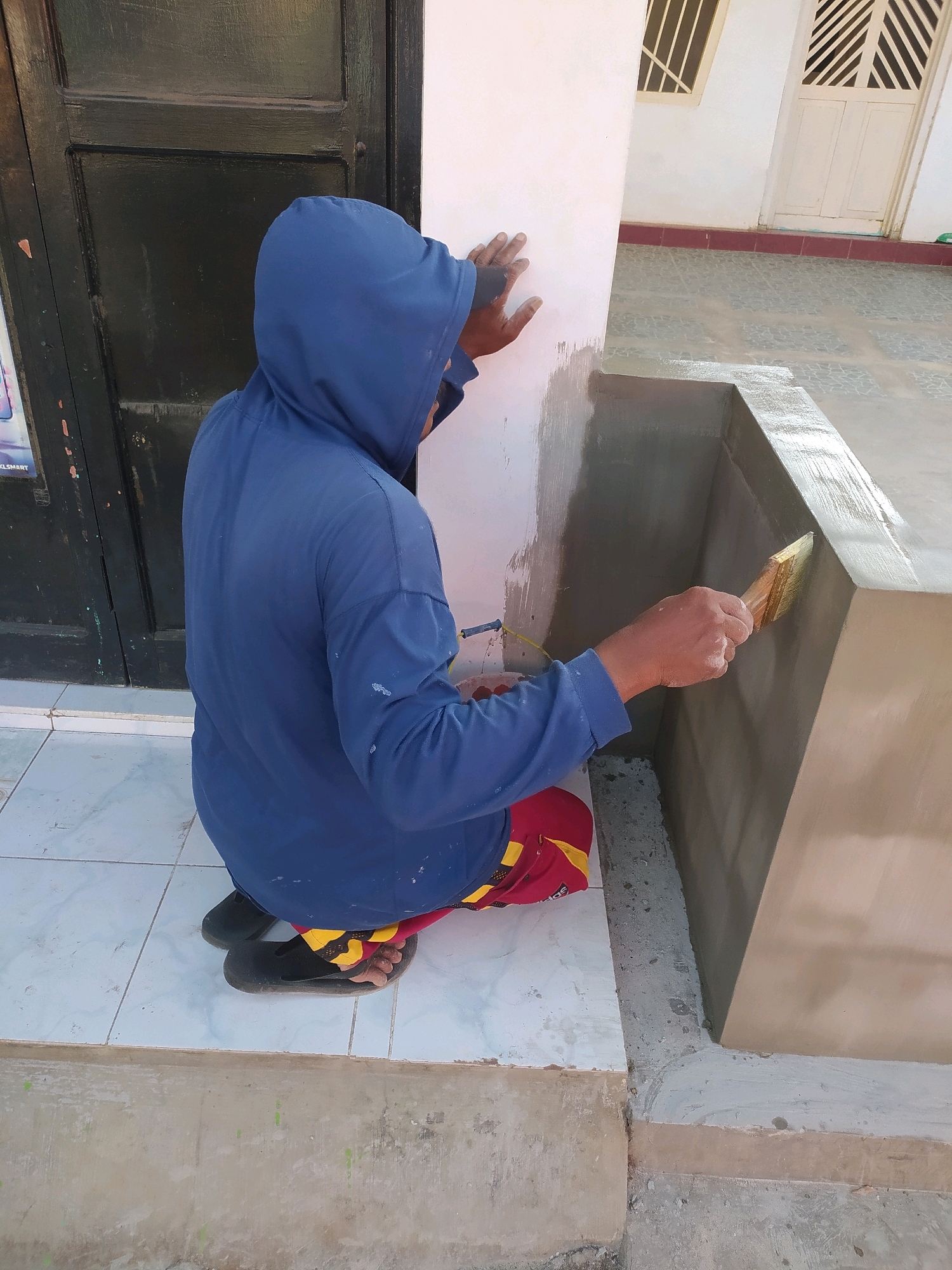An individual wearing a blue hooded sweatshirt and red and yellow shorts is crouched down, applying a gray coating to a concrete structure with a paintbrush. The person is positioned on a tiled floor in front of a dark green door. In the background, a white building with windows and a cream-colored door is visible. The scene appears to be outdoors, possibly in a residential area in Bantaran, Indonesia, given the architectural style and the warm lighting suggesting a daytime setting. The overall impression is of construction or renovation work in progress.
