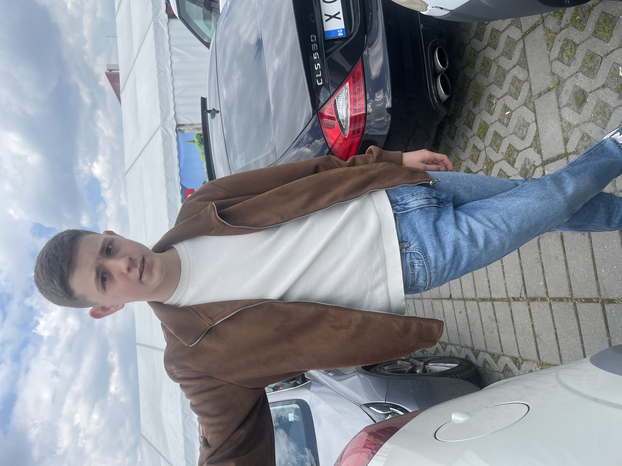 A young man stands among several cars. He wears a white, ribbed shirt, blue jeans, and a brown suede jacket. A black Mercedes CLS 550 is parked behind him, with a Bulgarian license plate beginning with "X". The cars are on a paved lot. The sky is overcast, and a large white tent structure is visible in the background. While the specific location within Stara Zagora cannot be determined from the image alone, the Bulgarian license plate indicates it is likely in Bulgaria.