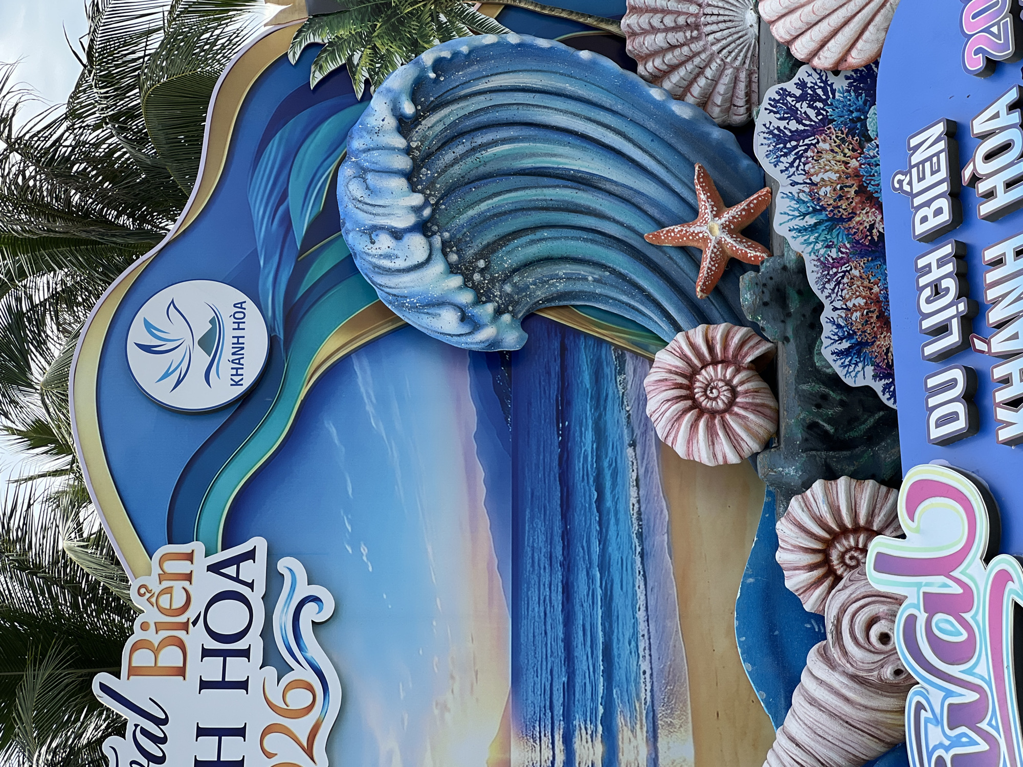 The image depicts a vibrant, decorative display for an event in Nha Trang, Vietnam. The central theme is the beach and the ocean, with a large, stylized blue wave prominently featured. Behind the wave is a backdrop that simulates a sunset over the sea and mountains. Various decorative elements, including large seashells, a starfish, and coral reefs, are placed in the foreground, adding to the beachy ambiance. The text on the display includes "Biển H HÒA 2026" and "DU LỊCH BIỂN KHÁNH HÒA 20". The logo "KHÁNH HÒA" with a bird and mountain motif is visible on a circular emblem. Palm fronds are visible at the top, suggesting an outdoor setting. There are no people visible in the image. The display appears to be set up on a blue surface, possibly a stage or a wall.