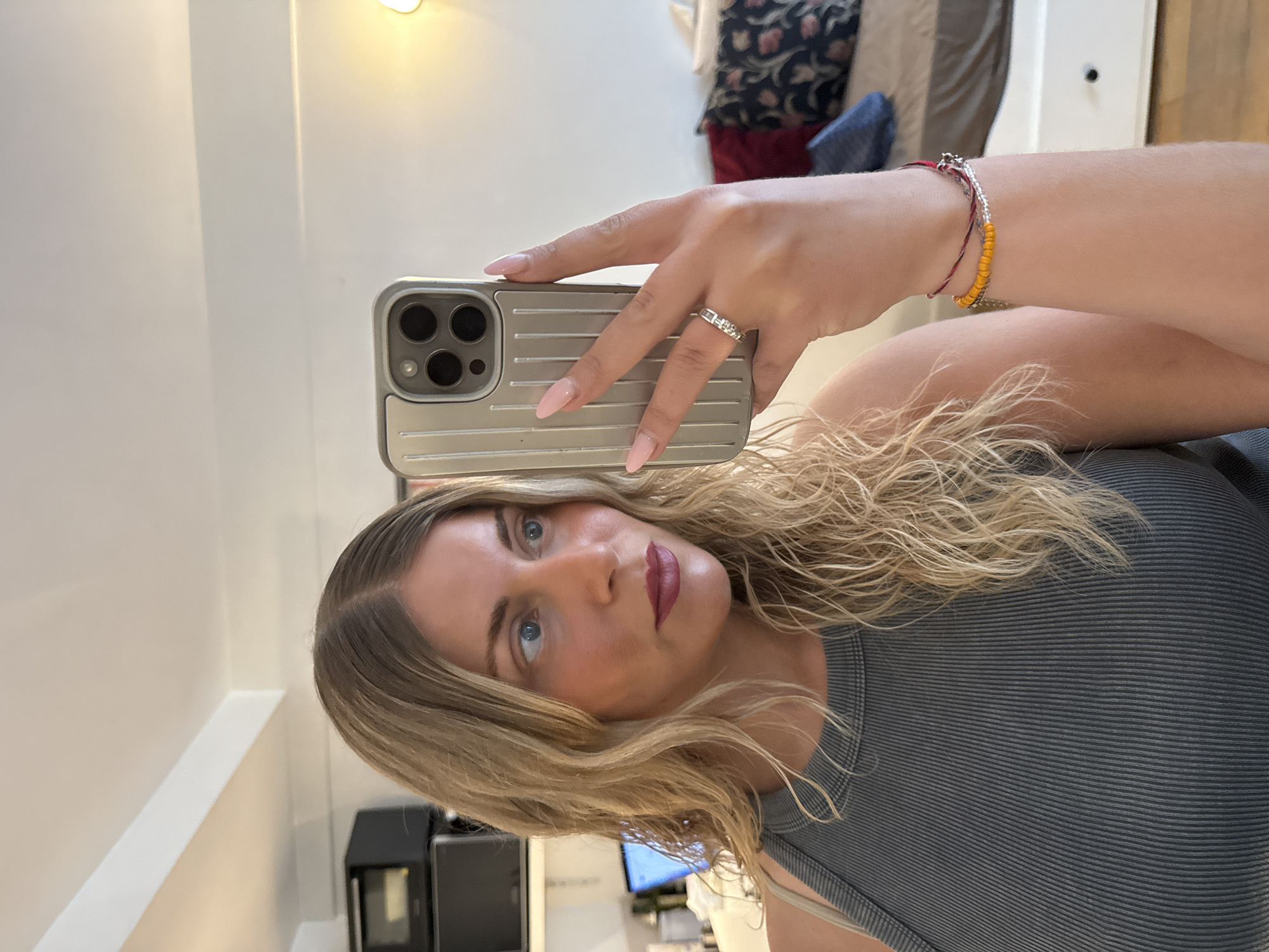 A woman with blonde, wavy hair is taking a selfie in a well-lit room. She is wearing a grey ribbed tank top and several bracelets on her left wrist. Her left hand is holding a silver iPhone with a ridged case, and she is wearing a ring on her ring finger. Her makeup includes dark lipstick and blush. In the background, there is a microwave on a shelf, a laptop with a blue screen, and some pillows on a bed. The room appears to be a bedroom or living space. There is no indication that this location is in Paris, France.
