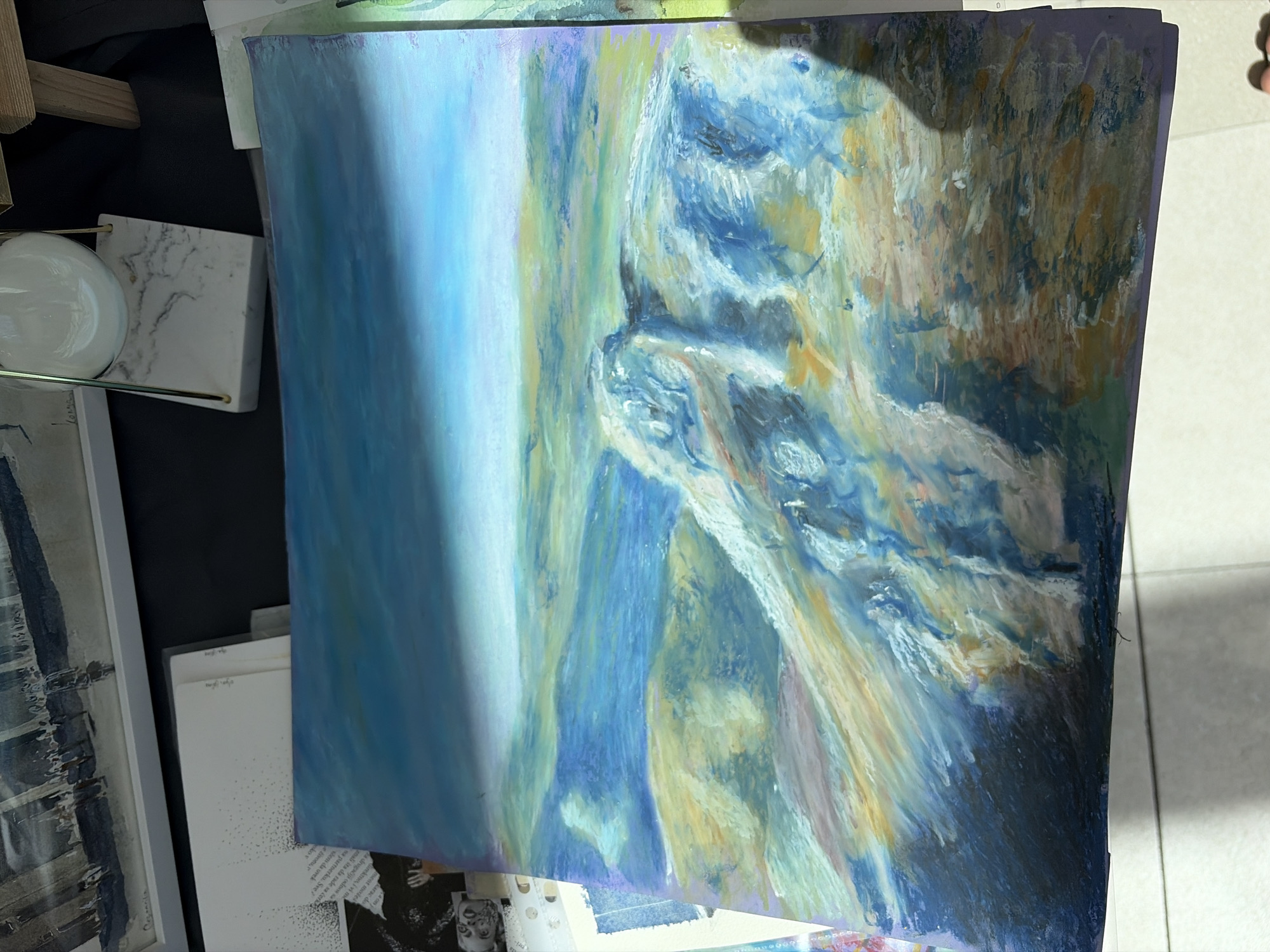 The image displays a pastel drawing of a landscape, possibly a mountainous or coastal region, with a prominent cliff face. The colors used are predominantly blues, greens, yellows, and whites, creating a sense of light and shadow. The sky is a gradient of deep blue to light blue, with a horizontal band of lighter color suggesting a horizon or a strong light source. The foreground features textured, rocky terrain rendered in bold strokes of blue and white, with hints of yellow and brown. The middle ground consists of rolling hills or slopes in shades of green and yellow. The drawing appears to be placed on a table, with other papers and objects partially visible in the background. No specific location in Budva, Montenegro is identifiable from the artwork itself.