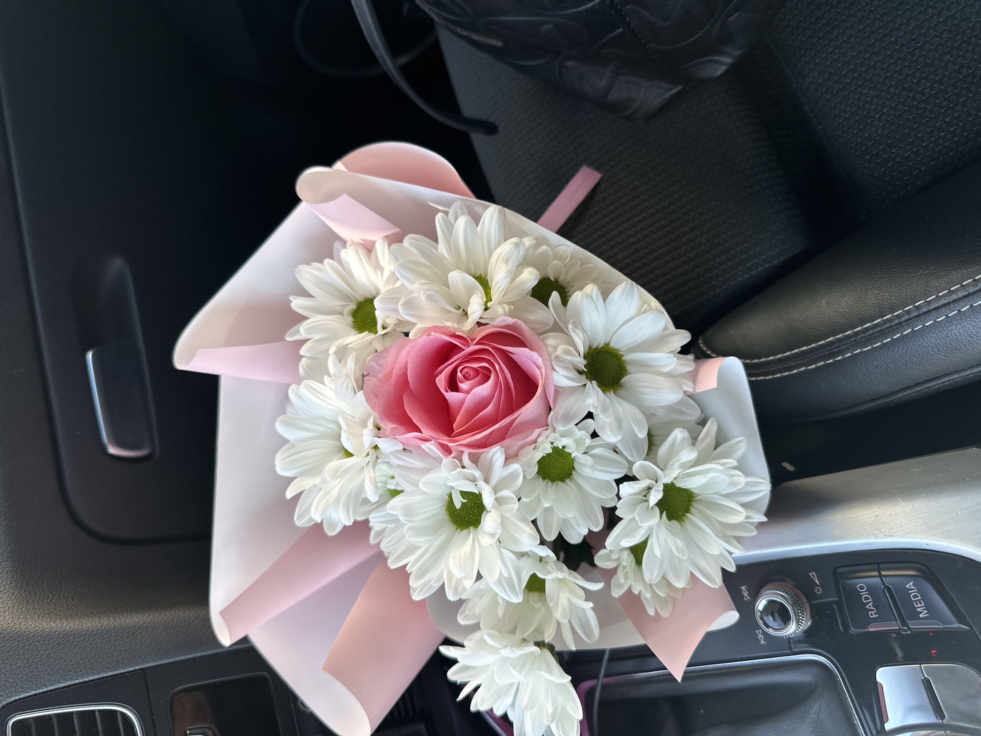 A bouquet of flowers is in a car. The bouquet contains white daisies and a pink rose. The flowers are wrapped in white and pink paper with a pink ribbon. The car's center console is visible, including the gear shift and radio controls. The seat is black leather with white stitching. The location is unspecified, but the question asked about the city of Sofia, Bulgaria. Therefore, it is implied the location is in Sofia, Bulgaria. No people are visible. The scene is indoors inside a car.