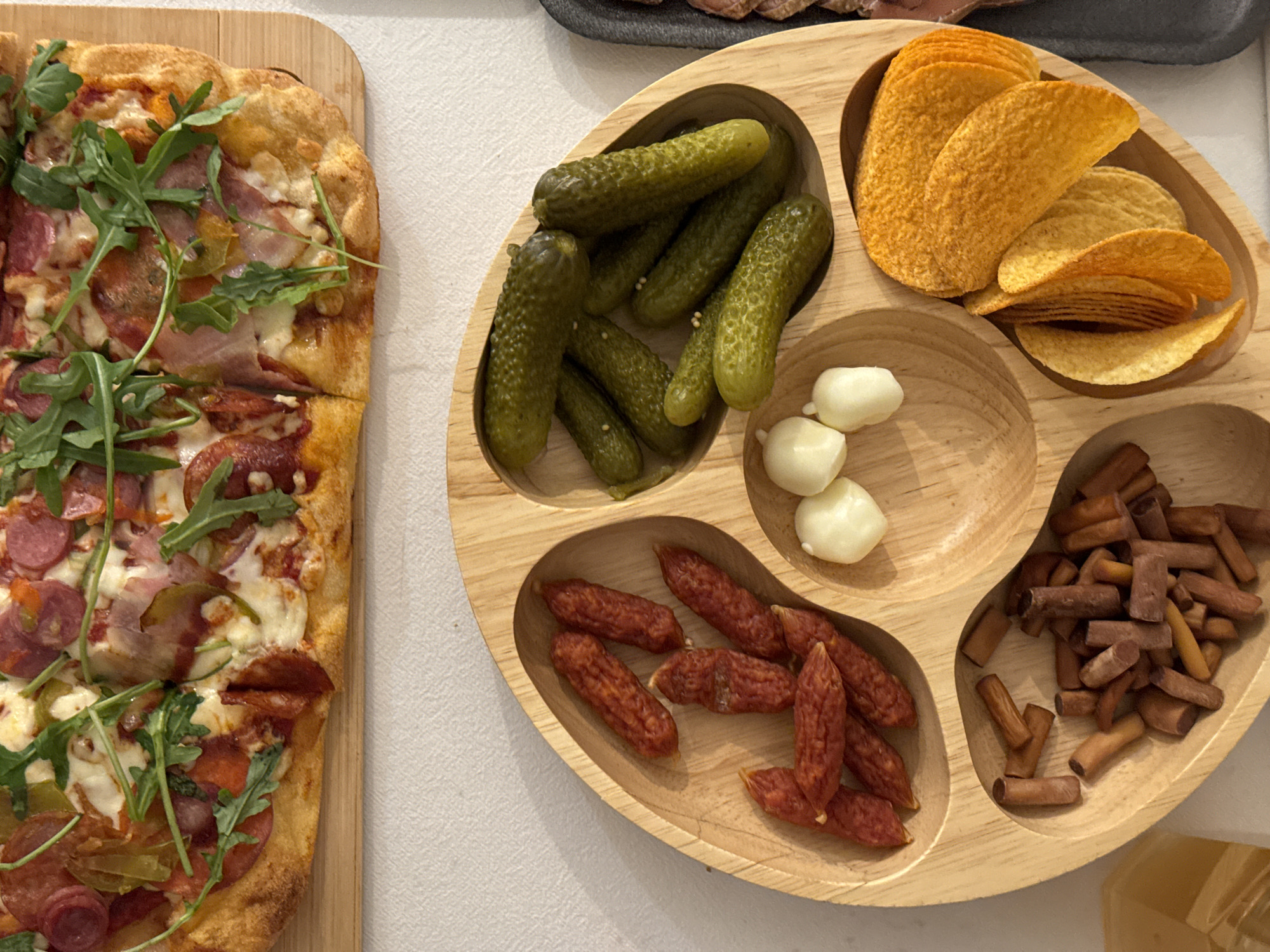 The image shows a spread of food on a table, likely for a casual gathering. On the left is a rectangular pizza on a wooden cutting board, topped with pepperoni, ham, cheese, and arugula. Adjacent to the pizza is a round, sectioned wooden platter. The platter contains various snacks: pickles, potato chips, small cheese balls, miniature sausages, and small breadsticks. The food is arranged on a light-colored surface, possibly a table cloth. The environment appears to be indoors, possibly a home or casual restaurant setting. There is no indication of this being in Akulovo (Gp Kubinka), Russia. No people are visible.