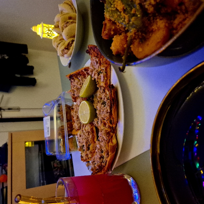A vibrant arrangement of food and drink sits on a table. A plate features what appears to be a savory pie topped with lemon slices. Nearby, there's a container of small bites. Another dish overflows with a mix of ingredients. A glass holds a red liquid, possibly juice or a cocktail, complete with a straw. A golden lantern and dishes enhance the scene, all set under colorful lighting.