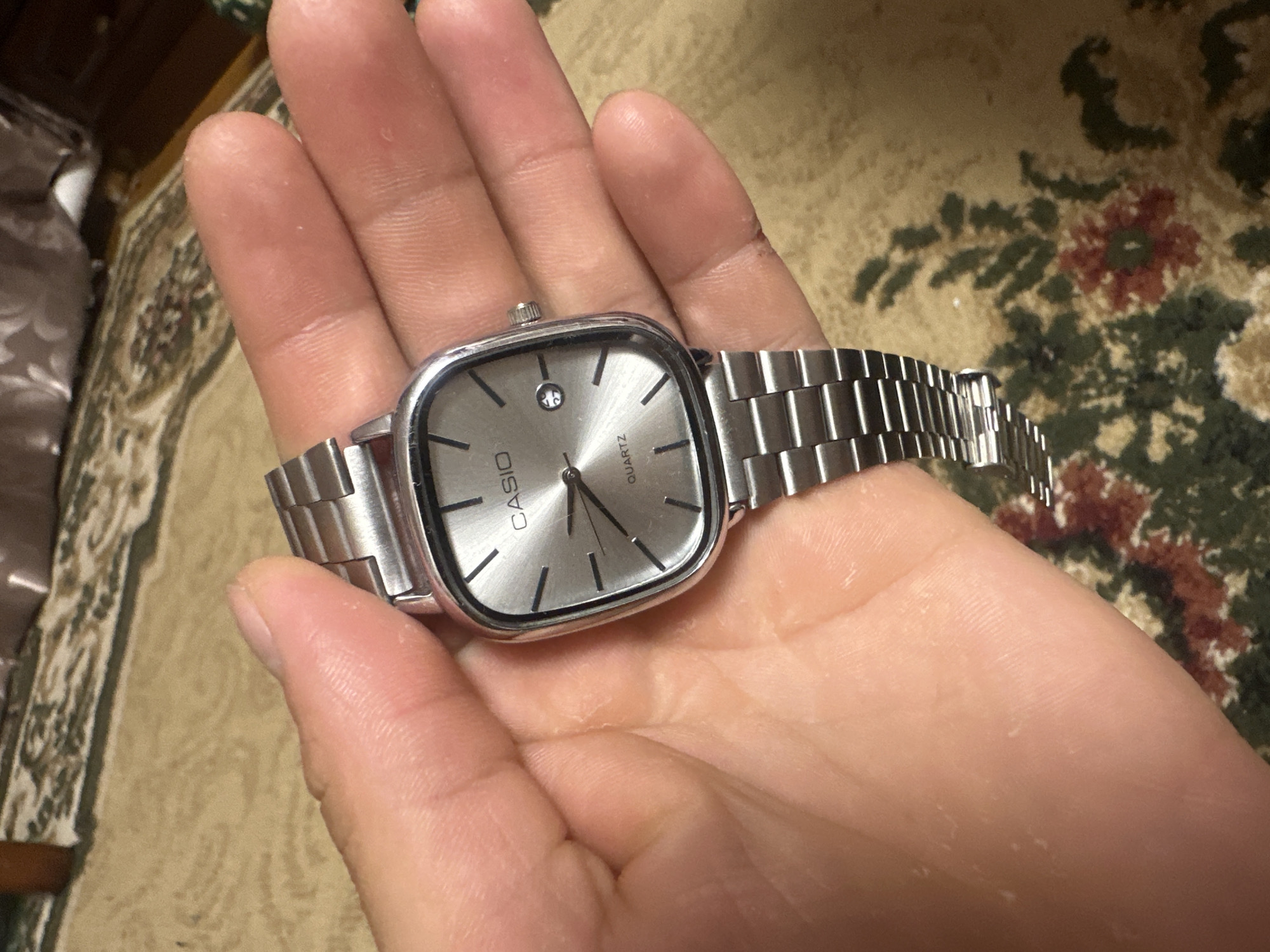The image shows a close-up view of a silver Casio watch with a cushion-shaped face being held in a person's hand. The watch has a metal band and a silver dial with black hour markers and hands. The word "CASIO" is visible at the top of the dial, and "QUARTZ" is visible below the center. The hand holding the watch appears to be light-skinned and has visible fingerprints. The background is a patterned carpet with floral designs in muted tones of beige, red, and green. The overall lighting suggests an indoor environment. No specific location in Загородный, Russia, can be identified from the visual information provided.