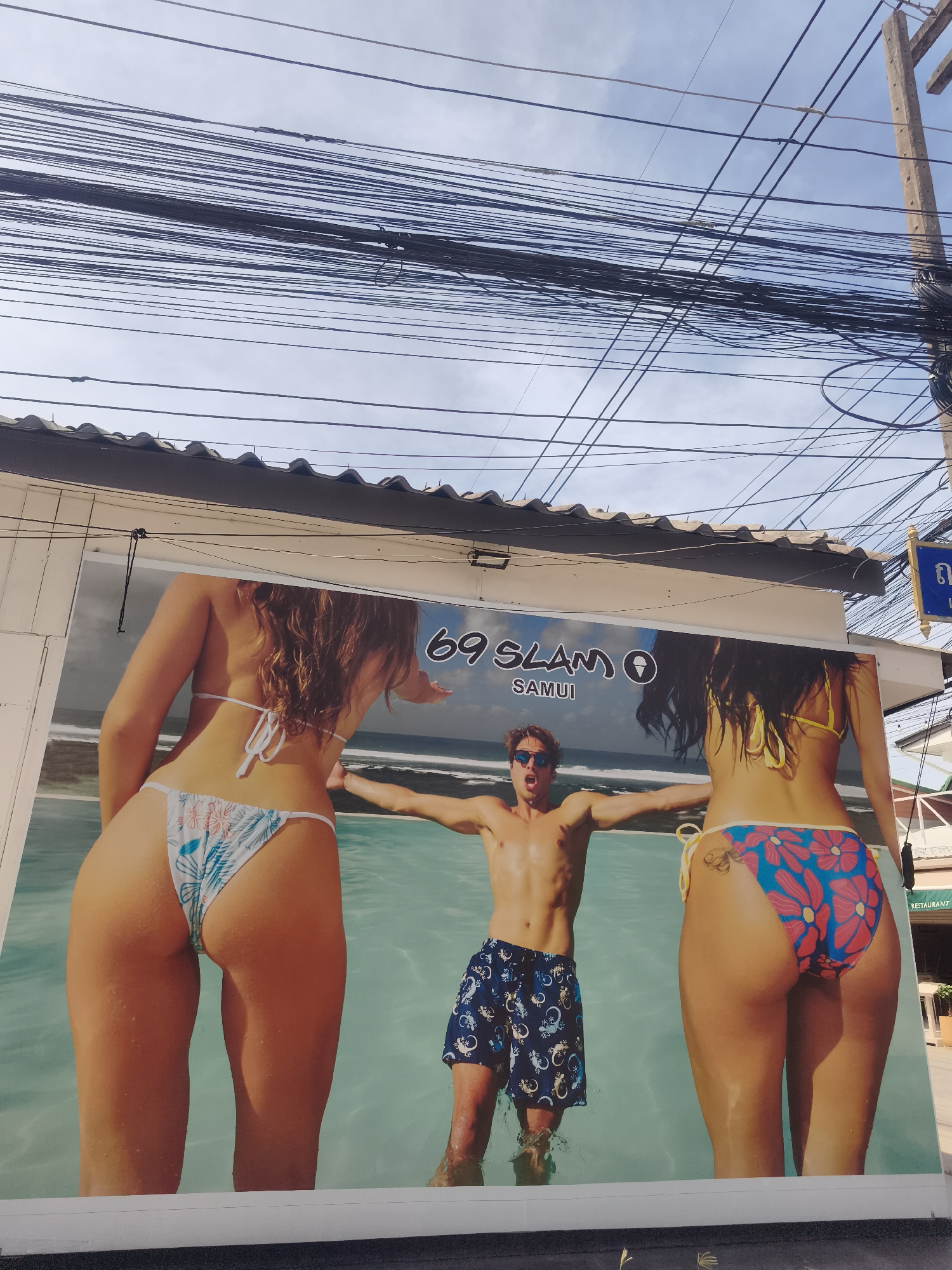 A large advertisement stands near a road in Baan Bo Phut, Thailand. The sign features a central male figure, shirtless and in swimming trunks, standing between two women whose rear ends are prominently displayed in bikinis. Overhead, a dense network of power lines tangles against a pale sky. A utility pole is visible on the right, and partial signage in Thai script appears. A hint of a building facade peeks out behind the sign, labeled 'Restaurant' on a vertical sign.