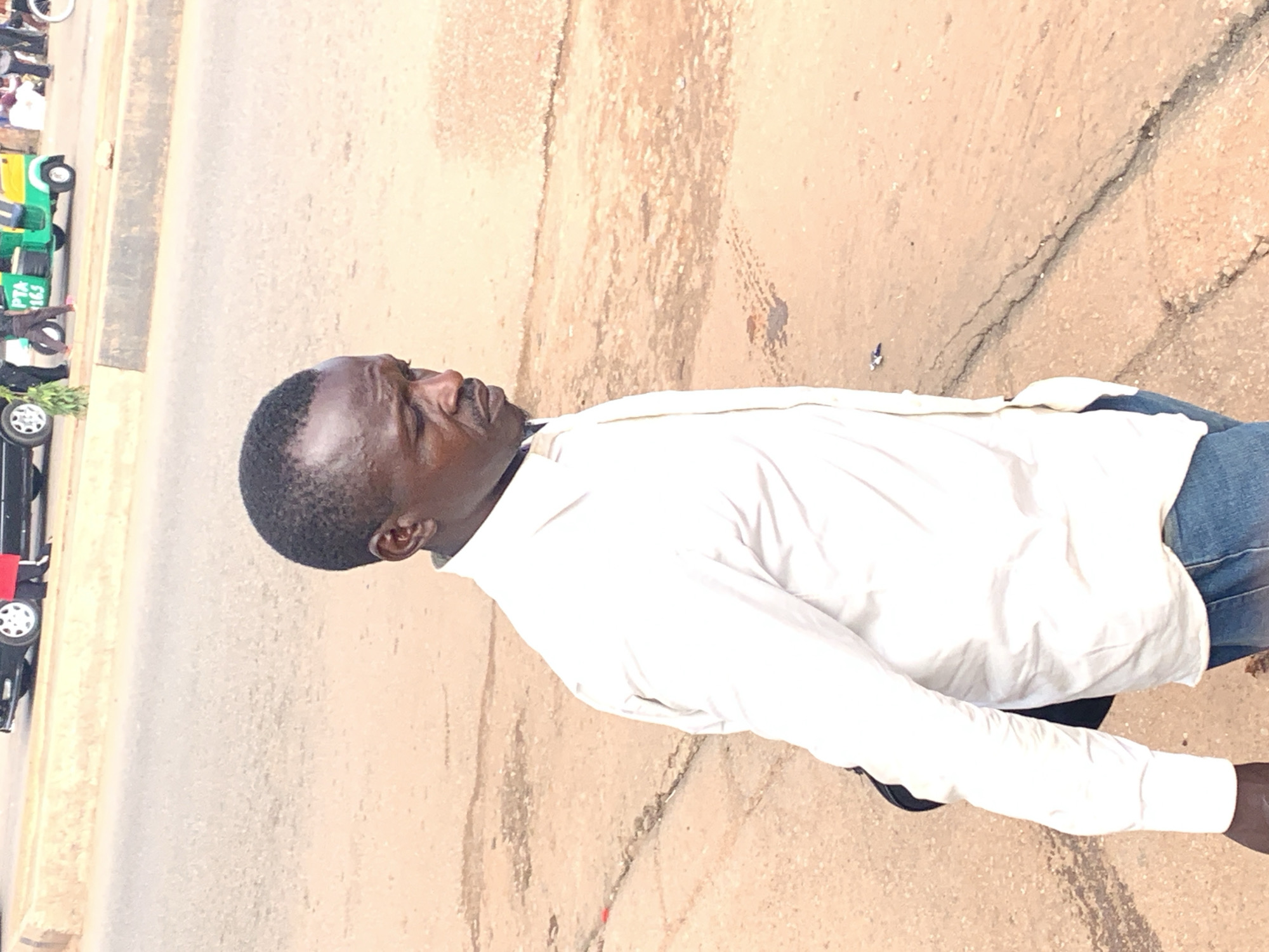 In Gura Top, Nigeria, a man with dark skin and short hair is captured in a medium shot. He is wearing a white button-down shirt, unbuttoned at the top, and blue denim jeans. A black bag is slung over his left shoulder. The man is looking down and to his right, his gaze directed away from the camera. He has a mustache and a goatee. The background shows a dusty, cracked asphalt road with a concrete barrier on the left side. In the distance, various vehicles are visible, including a green and yellow tricycle with the text "PTA 165" on its side, a black car, and other individuals walking or standing. The lighting suggests an outdoor setting during the daytime.