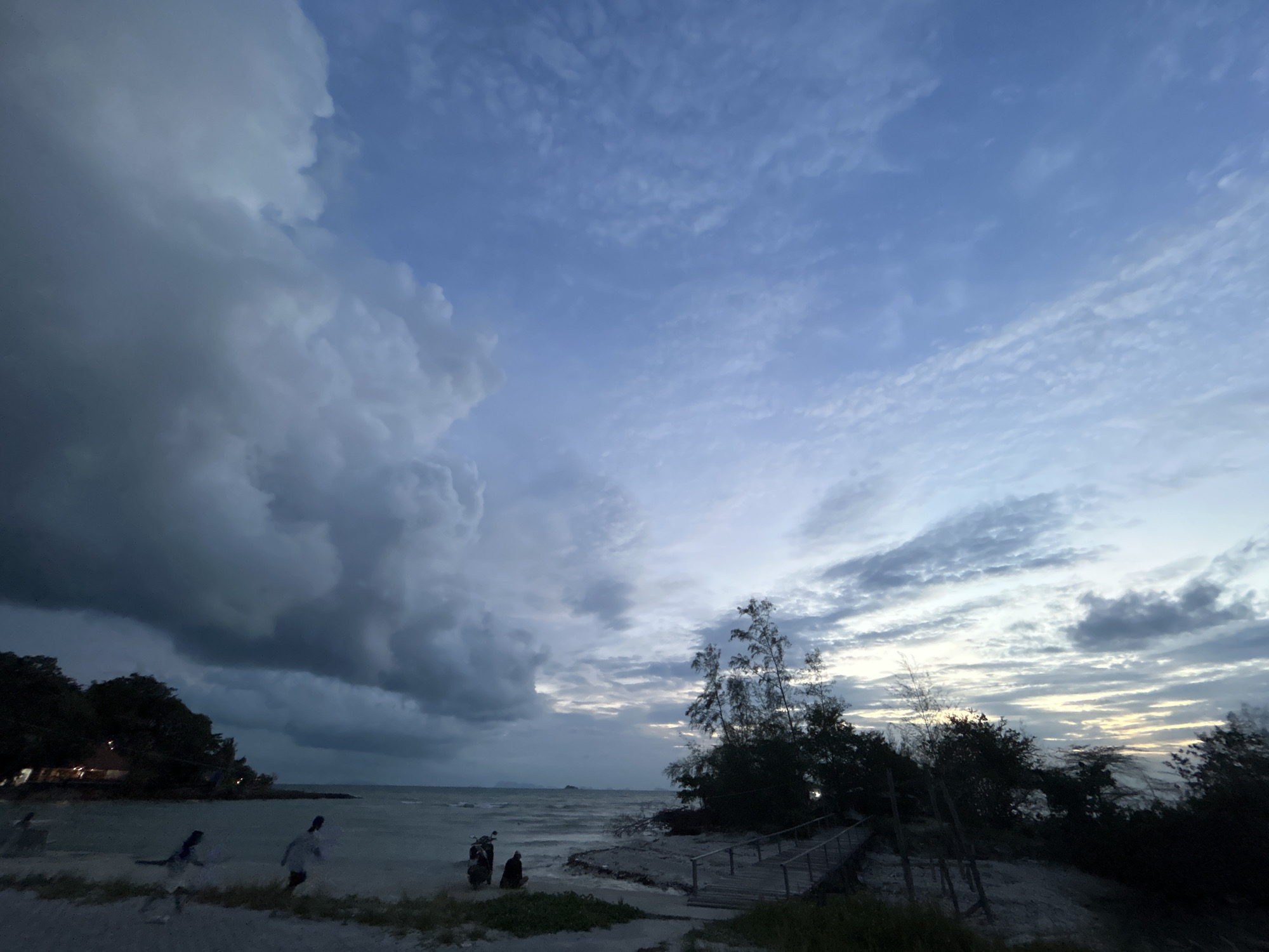 The image captures a dramatic sunset over a beach in the Ko Pha-ngan District, Thailand. The sky is a canvas of swirling clouds, with dark, ominous thunderclouds dominating the left side of the frame, contrasting with lighter, streaky clouds on the right, hinting at the fading sunlight. The sea appears choppy with moderate waves.
On the sandy shore, several figures are present. Two individuals, possibly women, are in motion on the left side of the beach, their forms silhouetted against the dimly lit landscape. Further towards the right, near the water's edge, two motorcycles are parked. Next to them, a person is seated, facing the water. A wooden walkway with railings leads up a slight incline on the right side of the beach, disappearing into a cluster of dark trees and vegetation. The overall atmosphere is moody and serene, indicative of the transition from day to night, with the approaching weather adding a sense of drama.