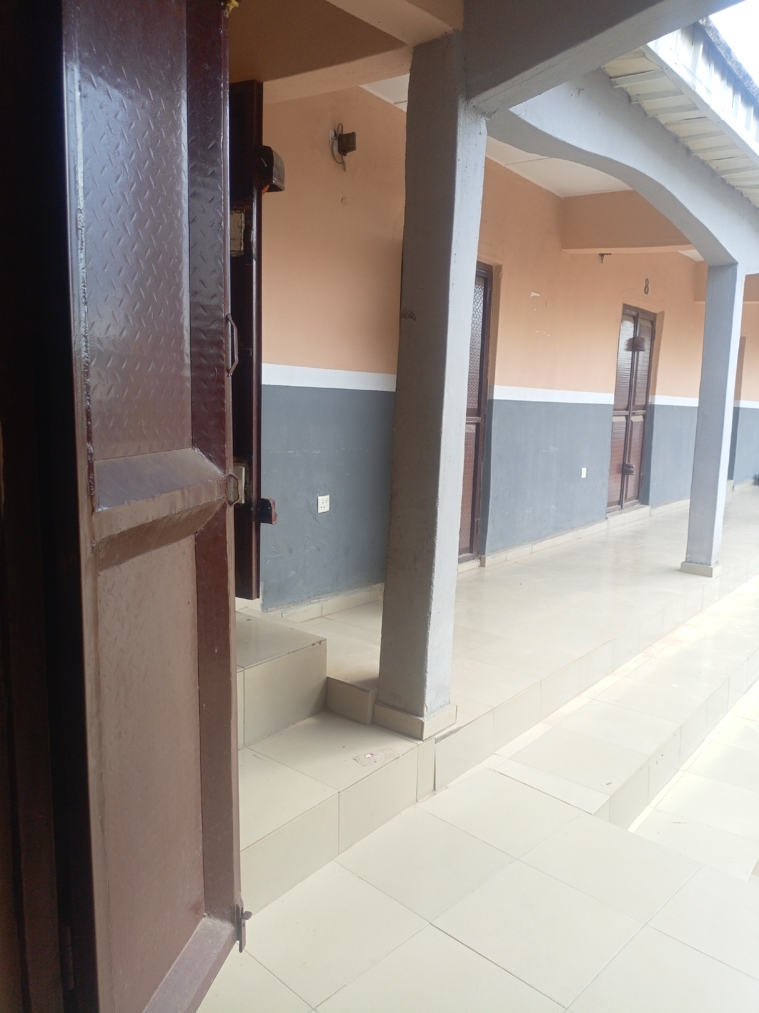 The image depicts an outdoor hallway or walkway, likely part of a residential or commercial building, with a tiled floor and a covered walkway supported by concrete pillars. The walls are painted in two horizontal sections, with the upper portion in a peach color and the lower portion in grey. Several doors are visible along the walkway, appearing to lead to individual units or rooms. One door in the foreground is partially open, revealing a metallic texture. Light enters from the right side of the image, suggesting an open-air environment. The setting appears to be in Jos, Nigeria, a city known for its moderate climate and a mix of modern and traditional architecture. The scene is devoid of people and activity, presenting a still and unoccupied environment.